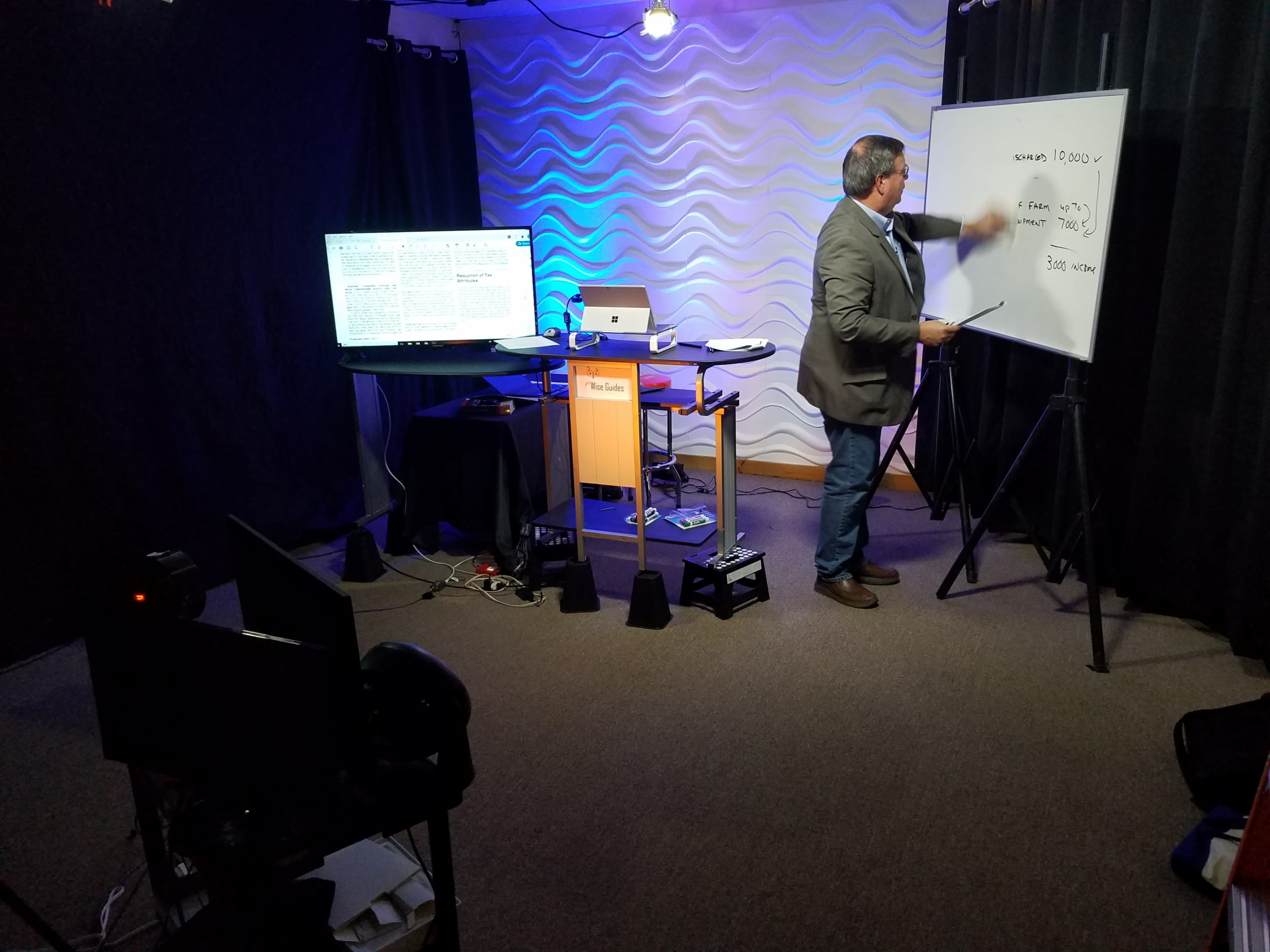 A presenter stands next to a whiteboard in a well-lit studio, with a monitor displaying text and a table containing equipment.