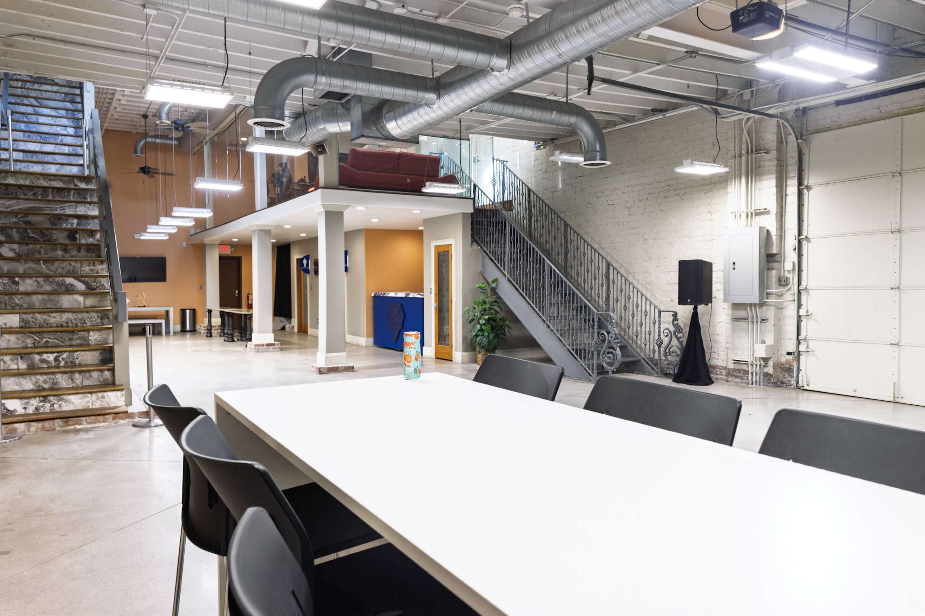 The image shows a spacious, industrial-style room with a long white table, black chairs, a staircase leading to an upper level, and a mix of seating areas in the background.