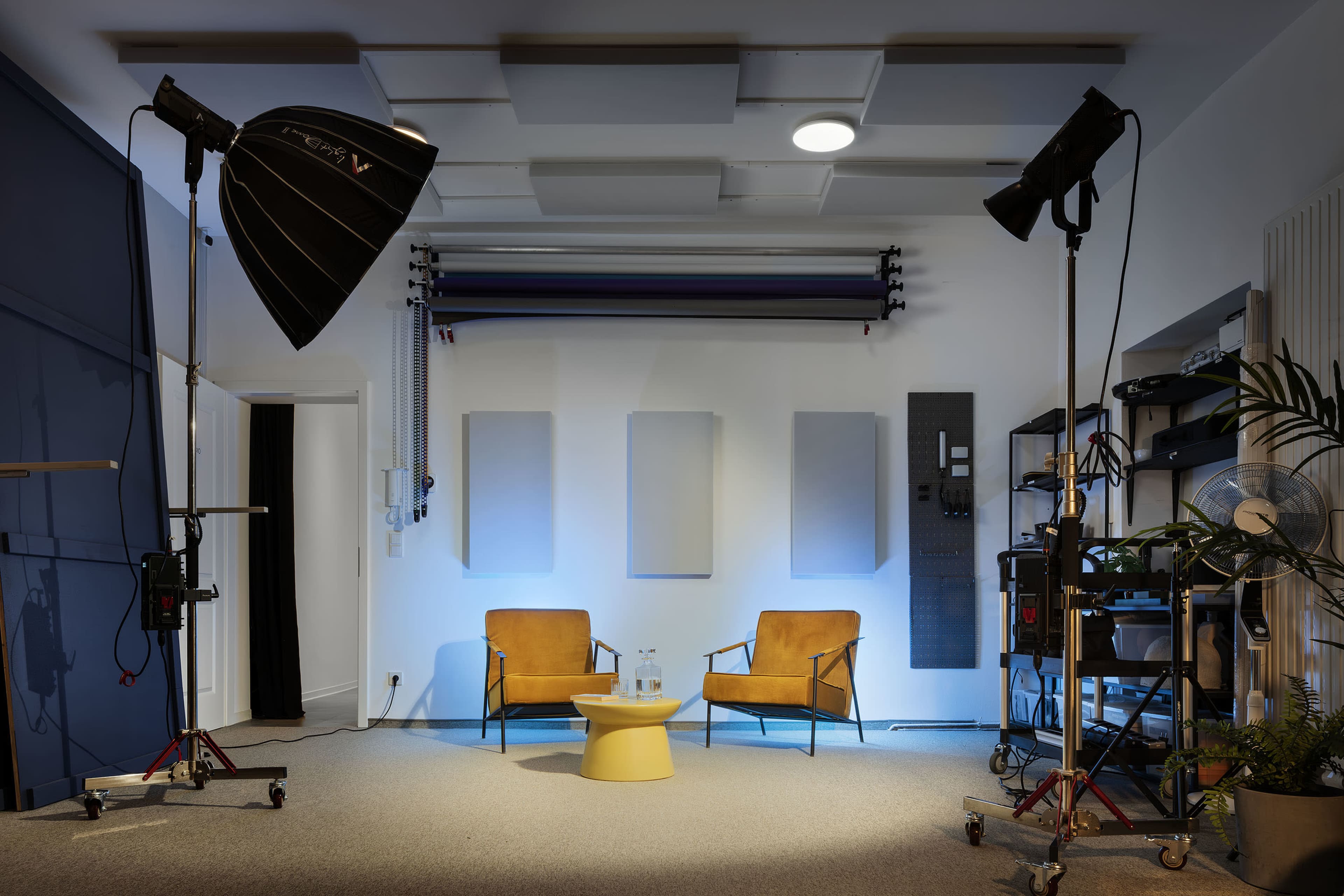 A well-lit studio space featuring two yellow chairs facing a small round table, with studio equipment and sound-absorbing panels in the background.