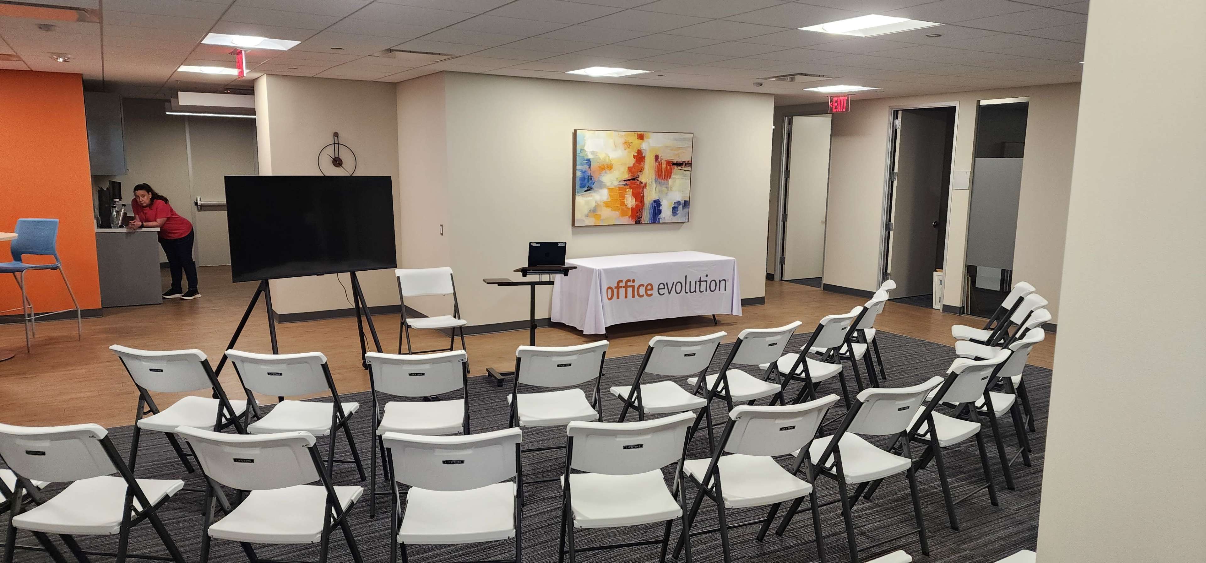 The image shows a room set up for an event, featuring rows of white chairs facing a presentation screen and a table with a branded tablecloth for "office evolution."