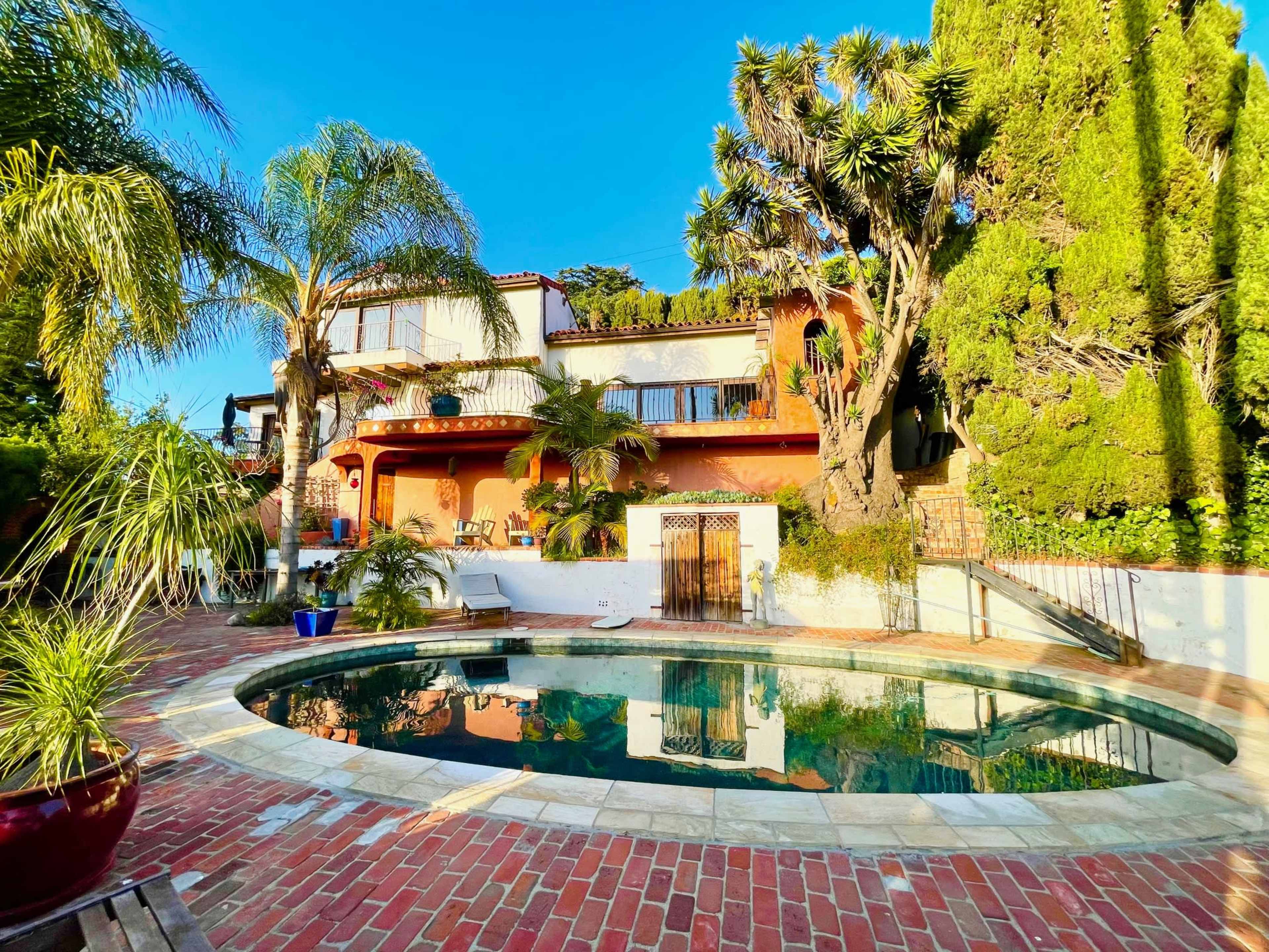 A landscaped backyard features a circular pool surrounded by brick paving and tropical plants, with a house visible in the background.