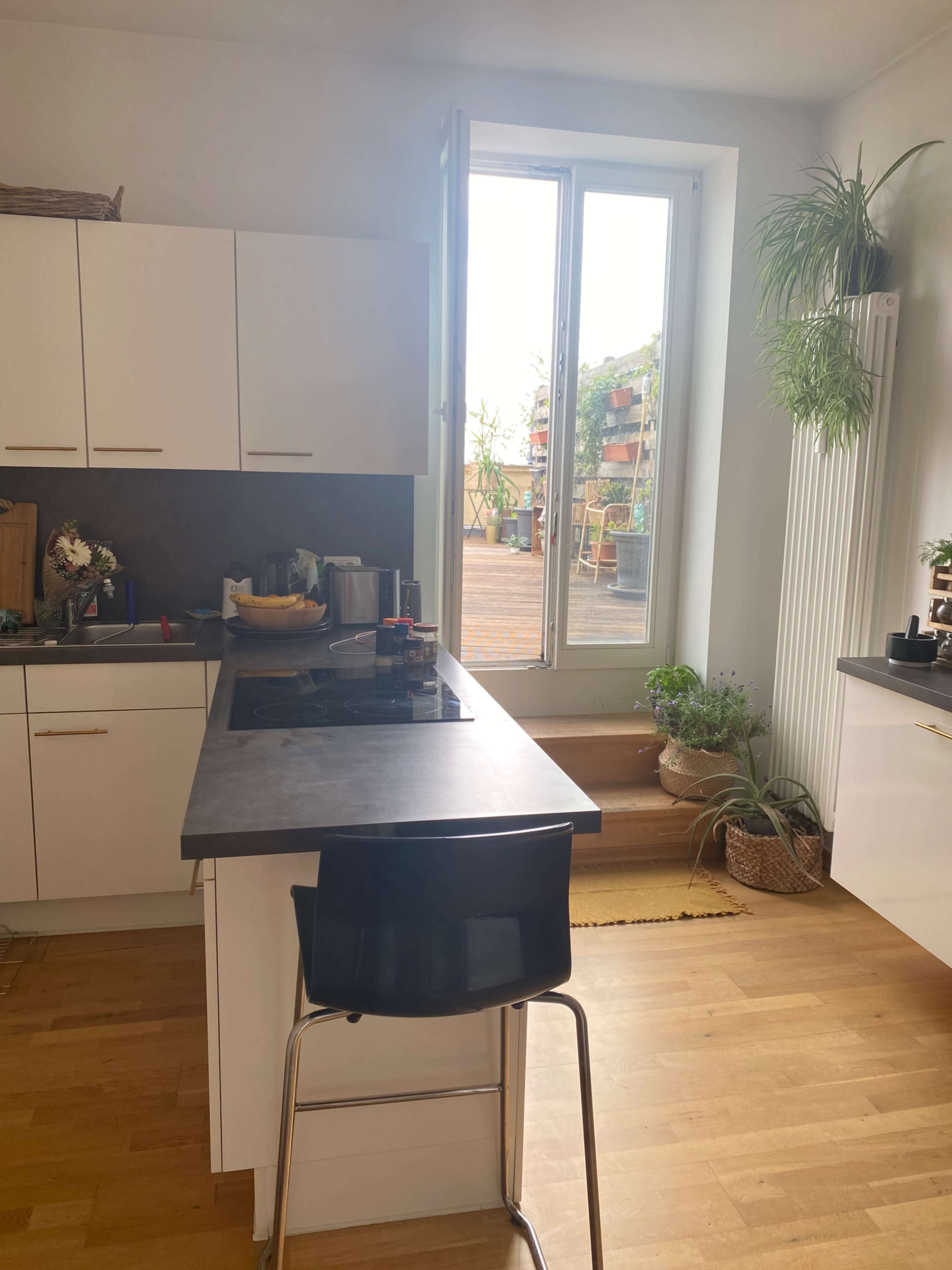 A modern kitchen with white cabinets, a gray countertop, and a large window overlooking a terrace filled with plants.