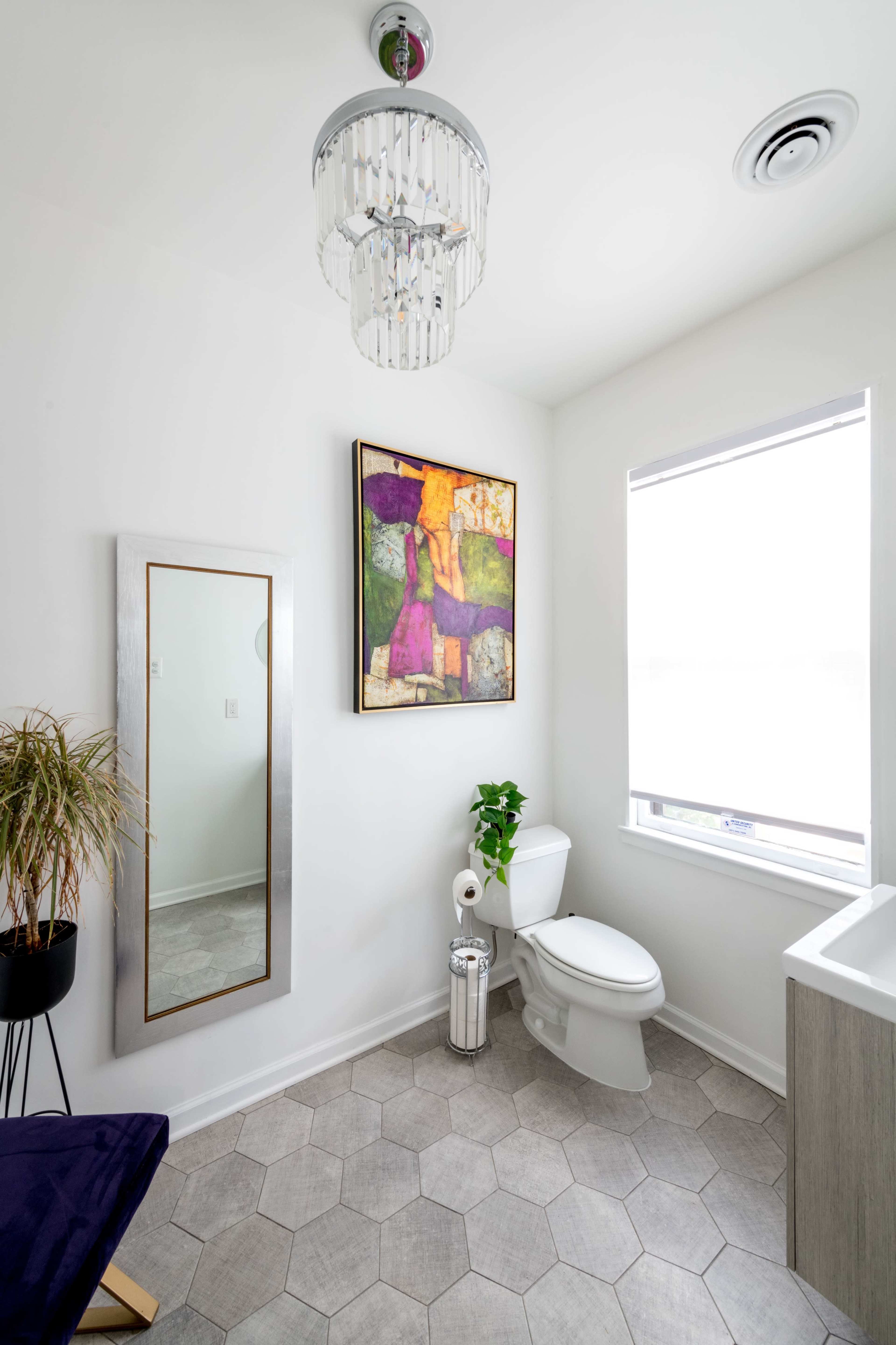 A modern bathroom featuring a chandelier, a large mirror, a colorful artwork on the wall, a toilet, a plant in a pot, and a sink.