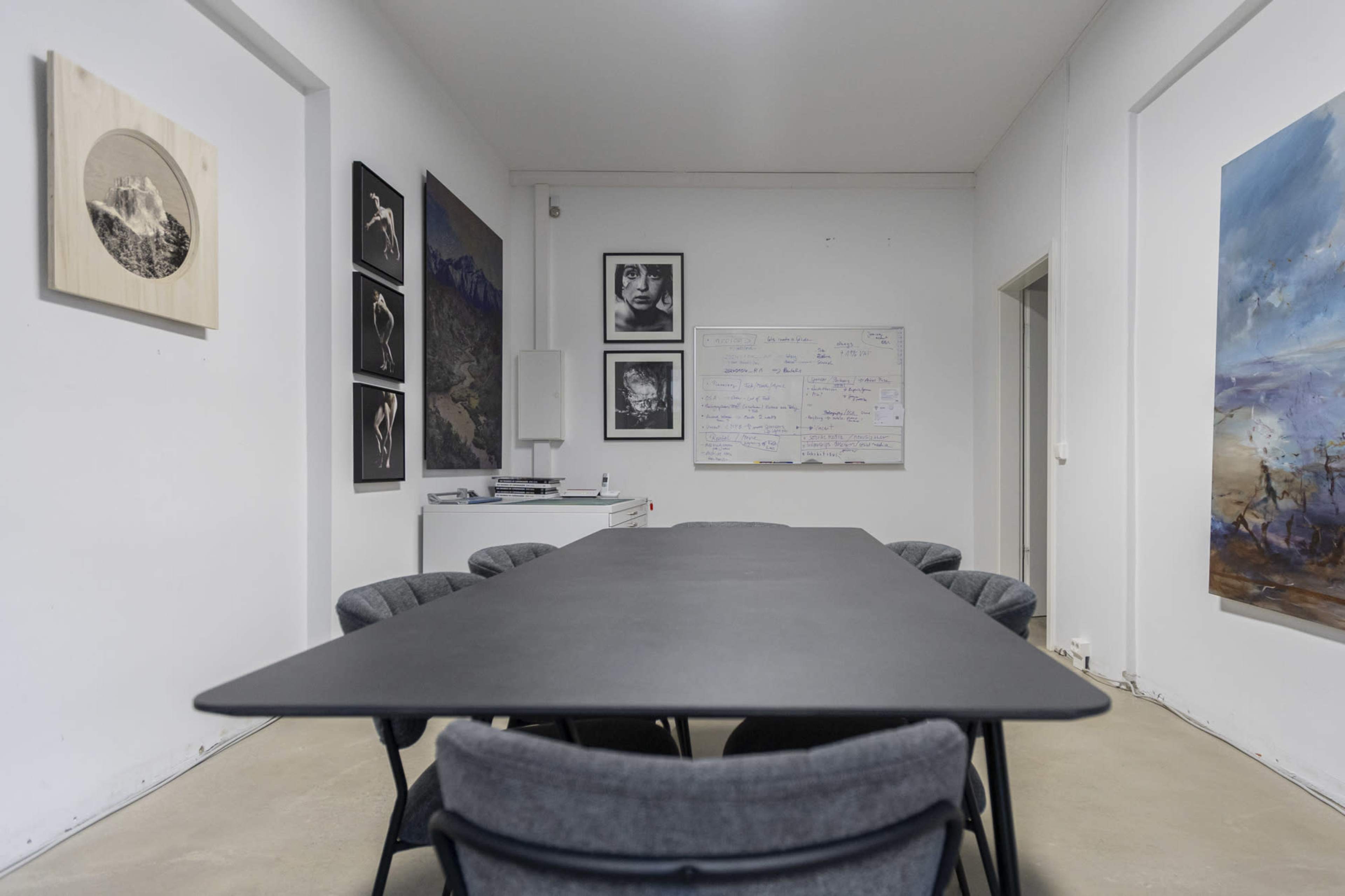A conference room features a long black table surrounded by gray chairs, with various artworks and a whiteboard on the walls.