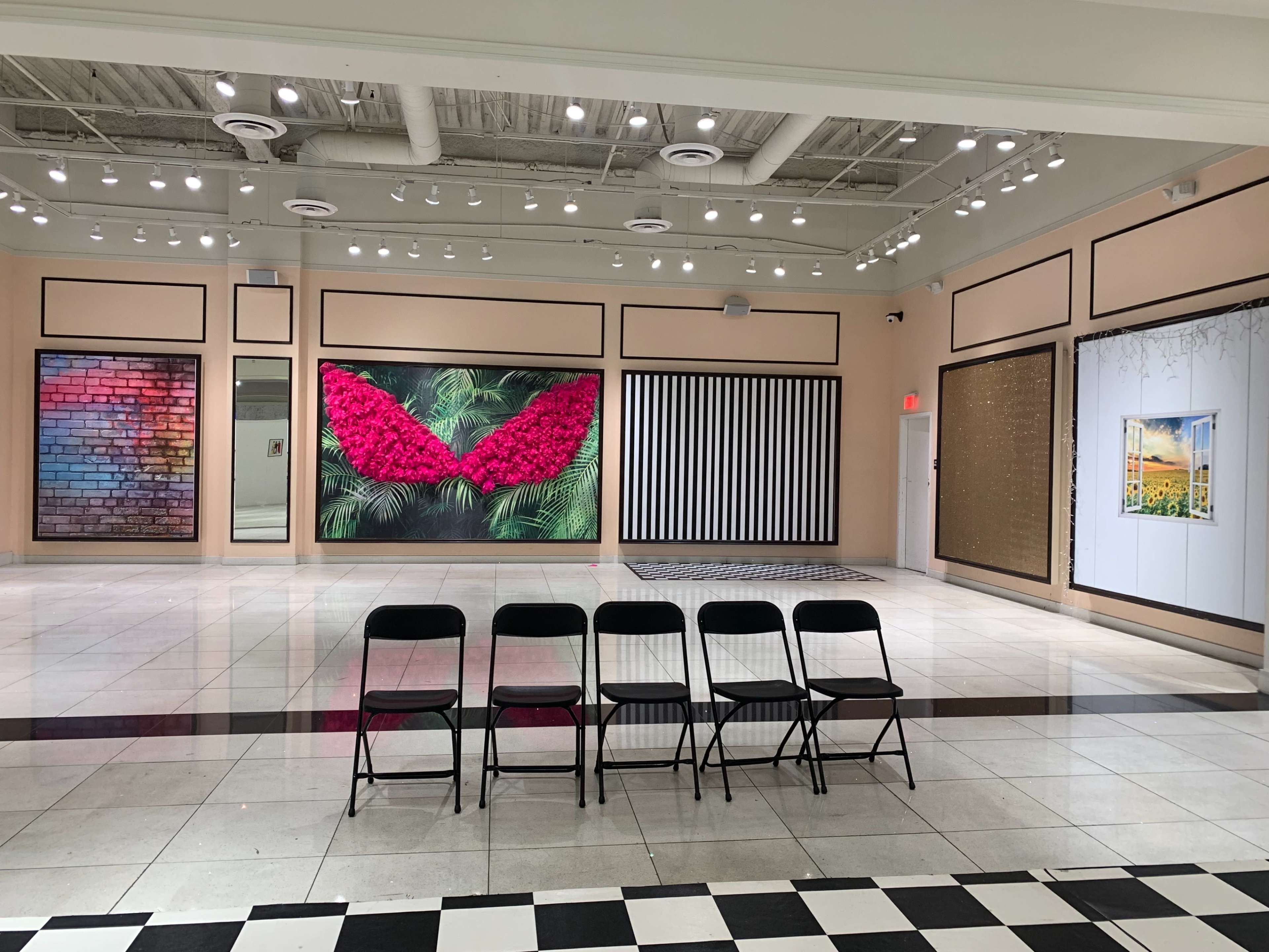 A gallery interior features several large artworks on the walls, including floral and textured designs, with a row of black chairs positioned in front.