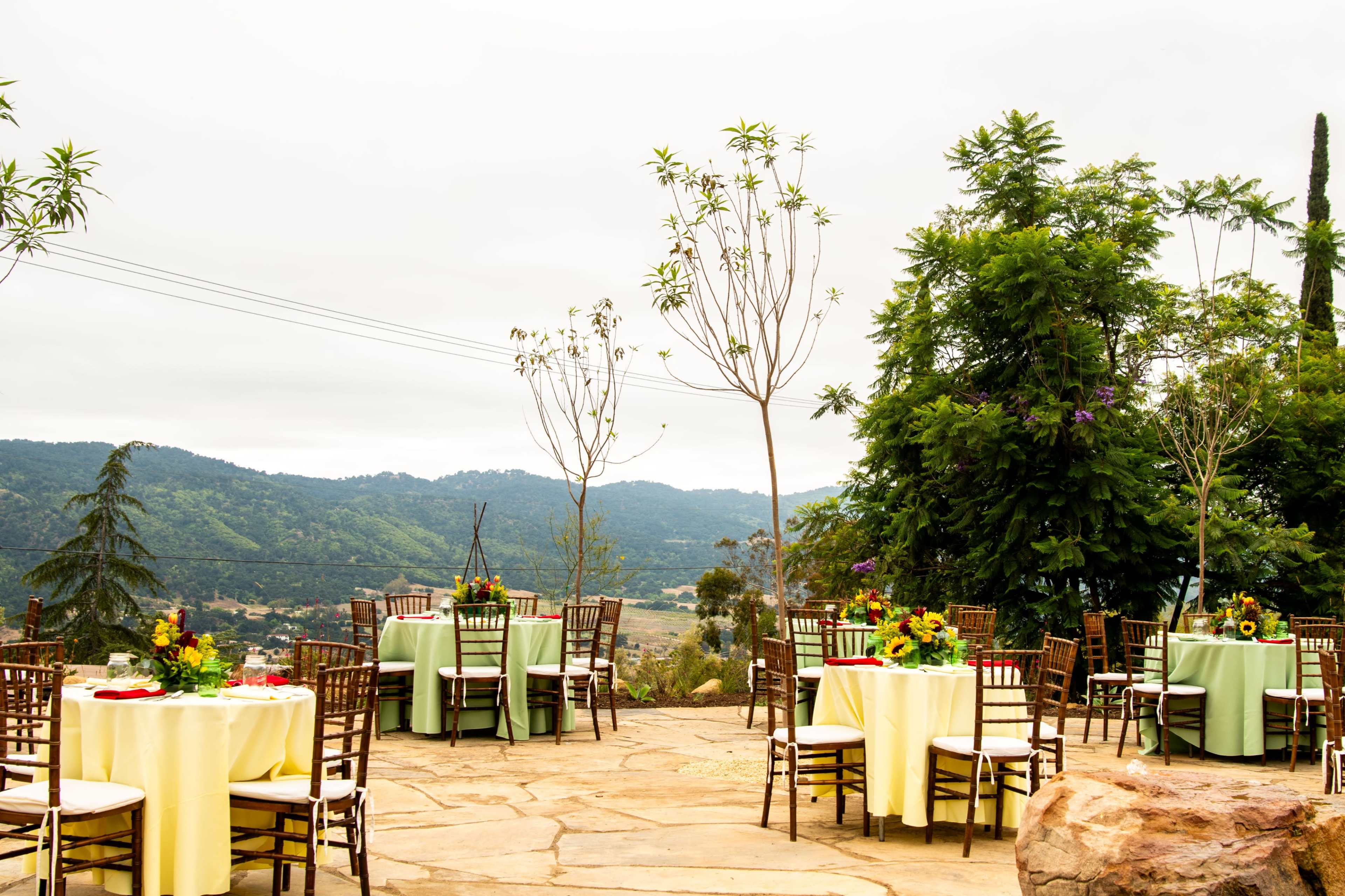 The scene shows an outdoor setting with several round tables covered in yellow and white tablecloths, surrounded by chairs and greenery, against a backdrop of hills and a cloudy sky.