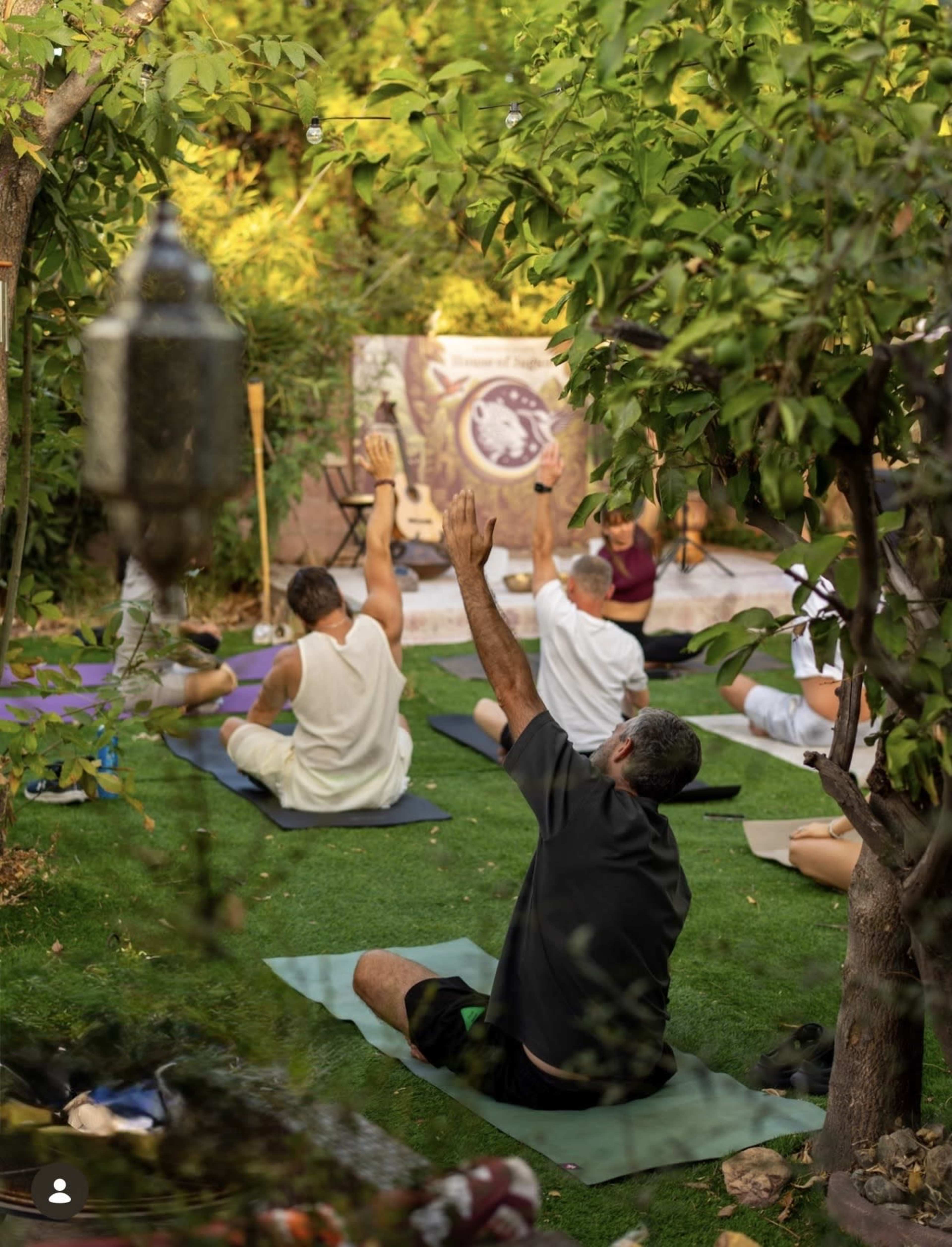 A group of people practices yoga outdoors in a lush garden, with some participants raising their arms in various poses.
