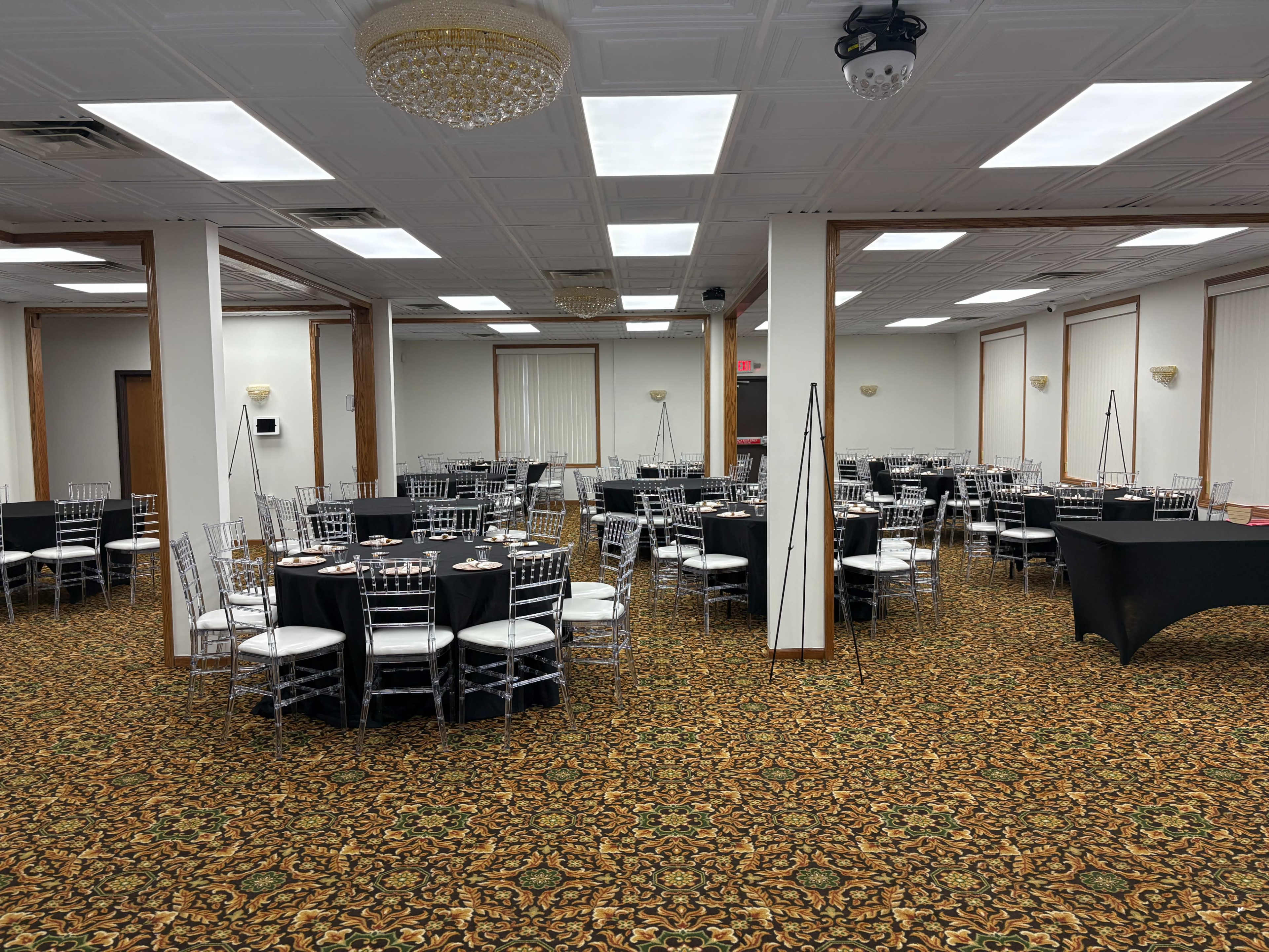 The image shows a spacious event hall with round tables, silver chiavari chairs, and a patterned carpet, arranged for a gathering or banquet.