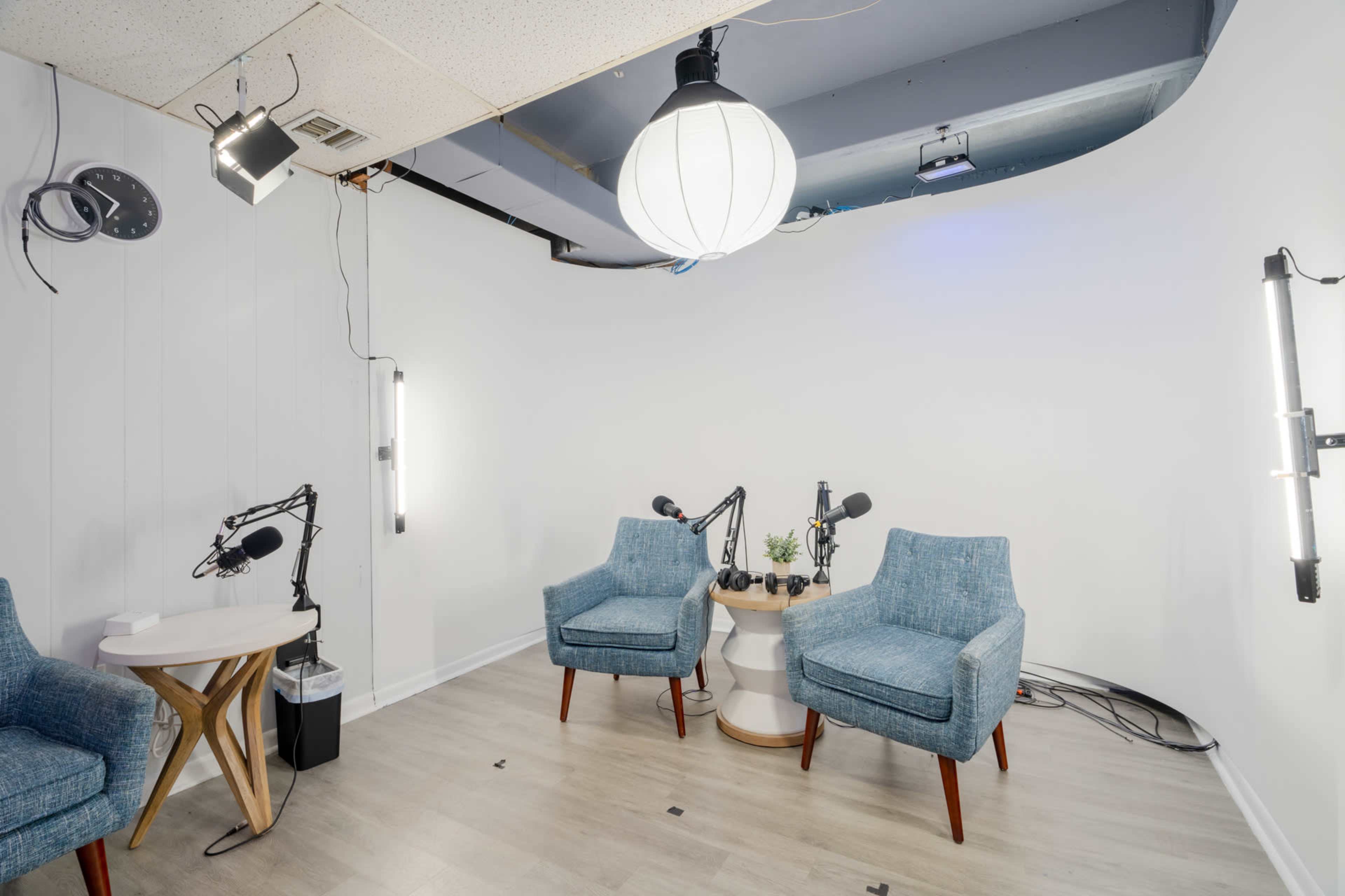 A minimalistic recording studio with two blue chairs, a small table, and microphones set up, illuminated by pendant lights.