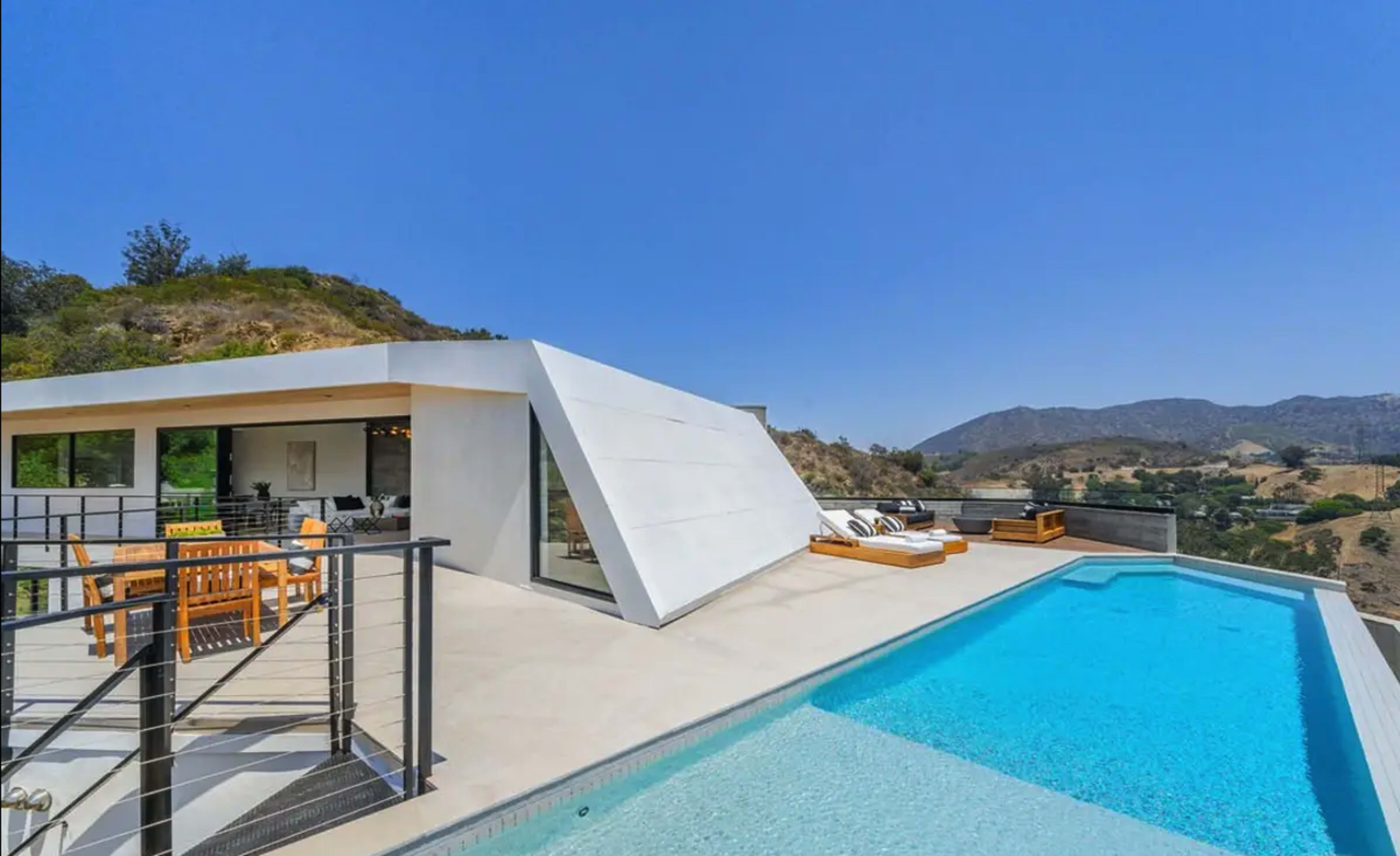 A modern, white geometric home overlooks a swimming pool and hillside landscape under a clear blue sky.