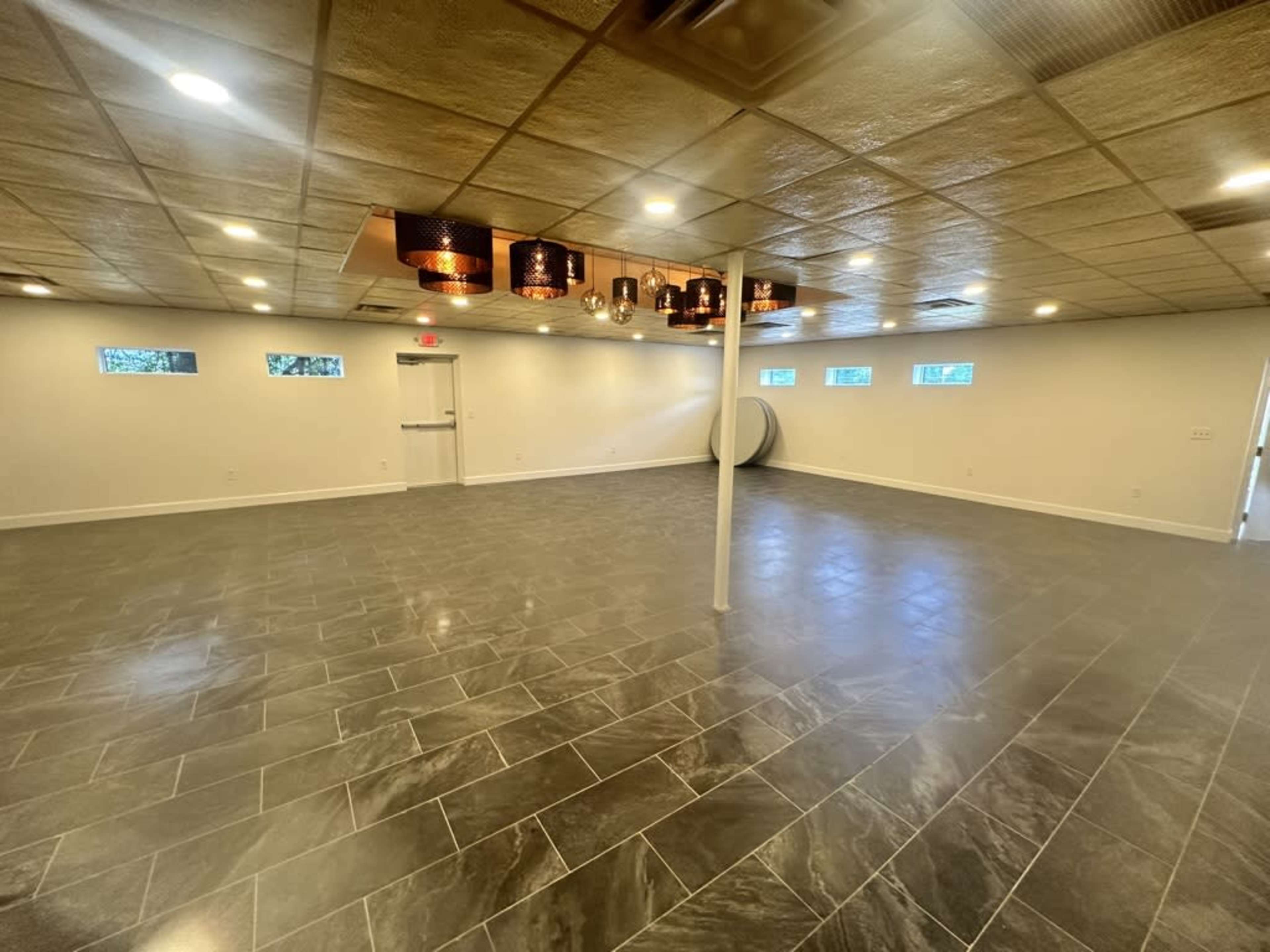 The image shows a spacious, empty interior room with gray tiled flooring and modern light fixtures hanging from the ceiling.