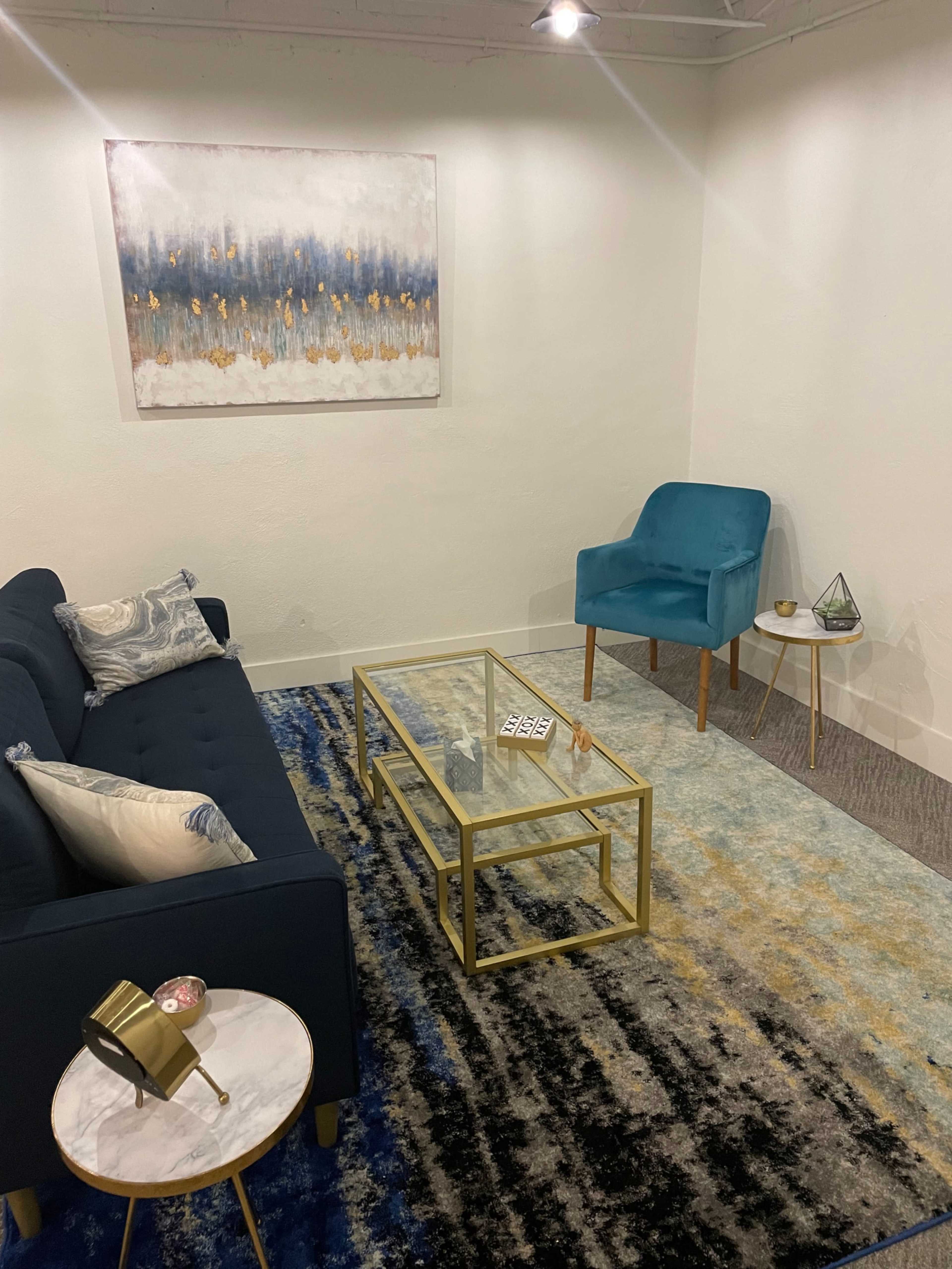 The image shows a small, modern living room featuring a blue couch, a glass coffee table, an accent chair, and a decorative area rug.