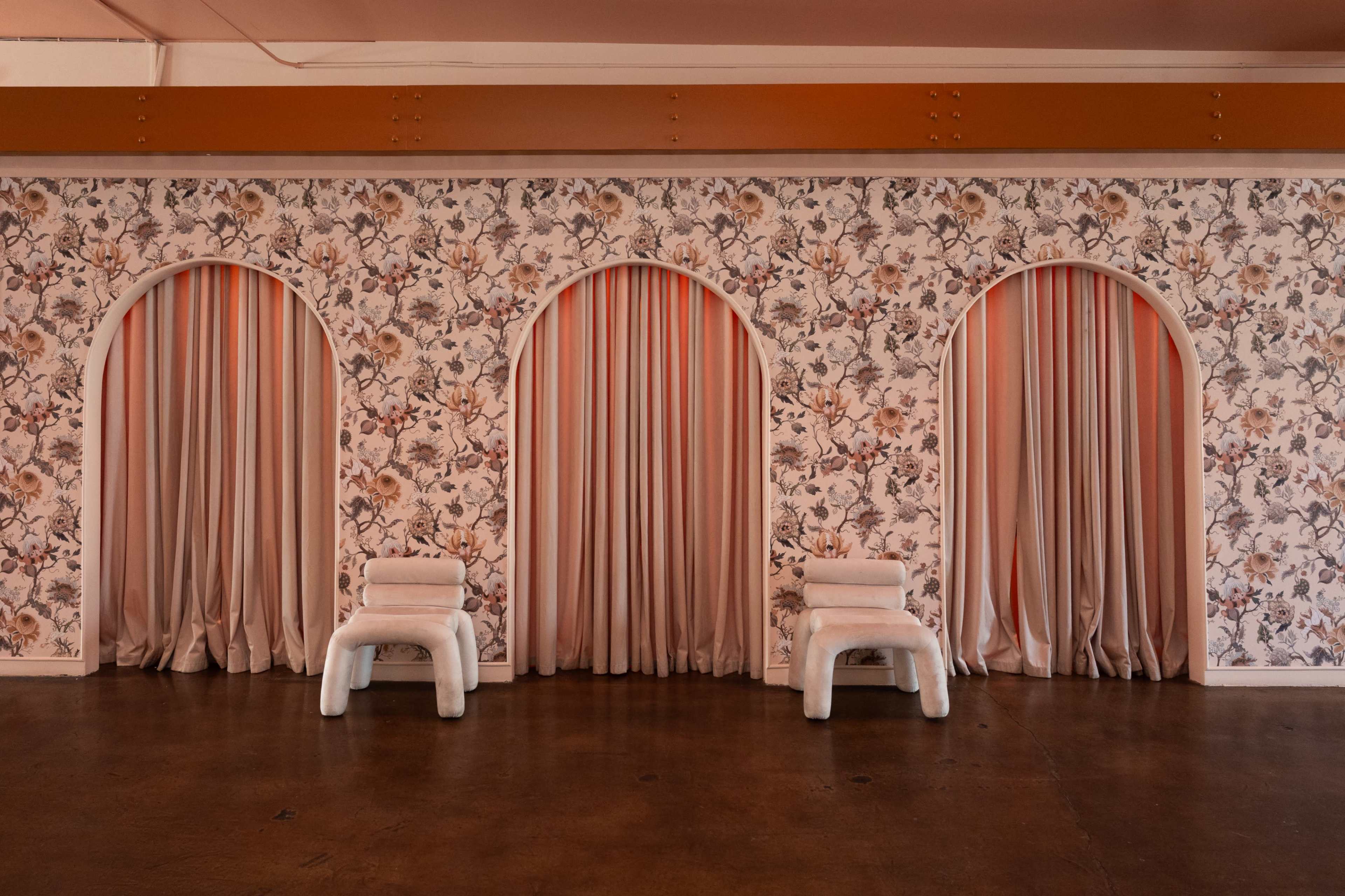 The image shows a room with a floral-patterned wallpaper, featuring three arched openings with pink curtains and two white, minimalist chairs placed in front.