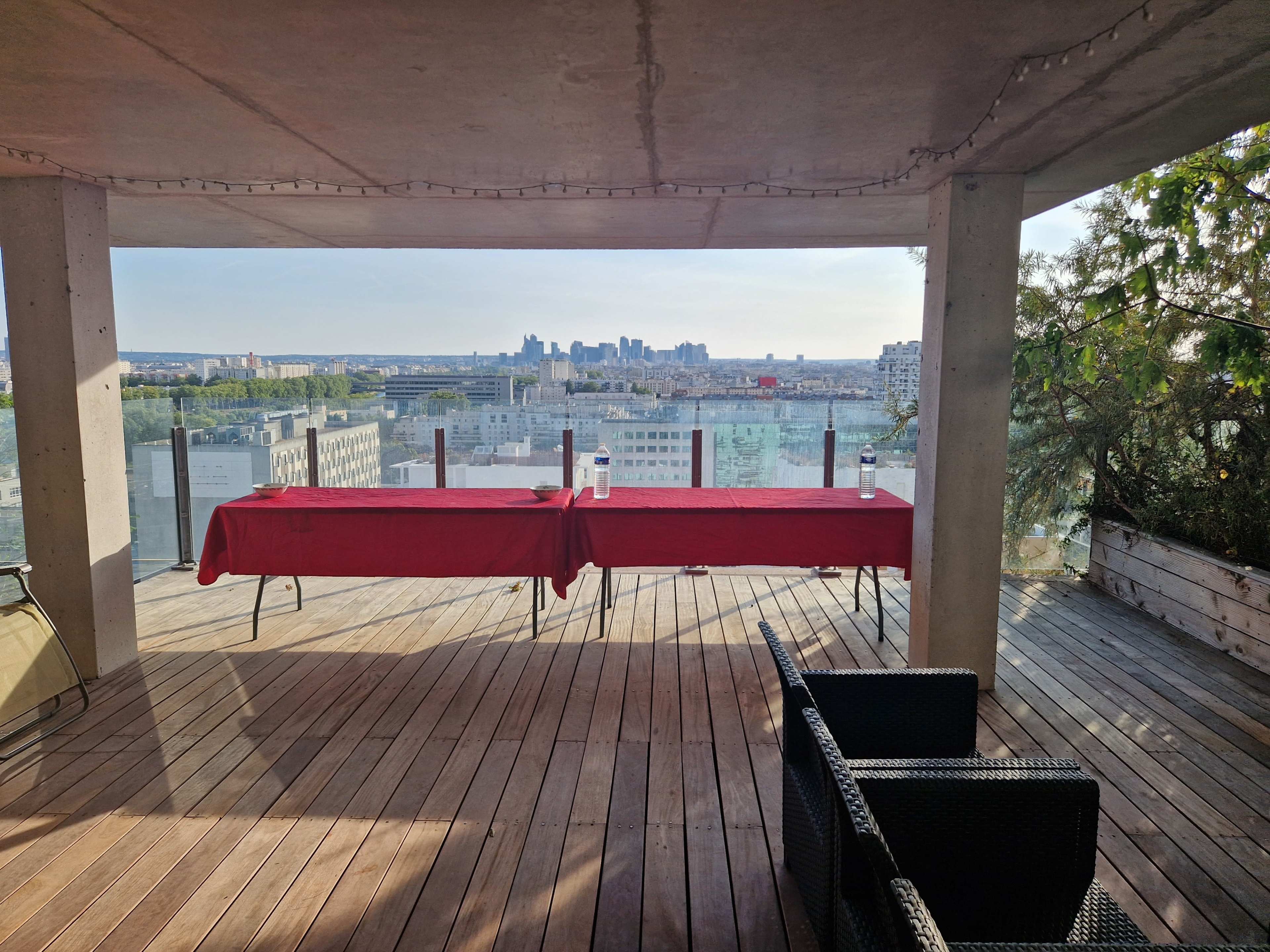 A spacious terrace features two red tables overlooking a city skyline, with seating arrangements in the foreground.