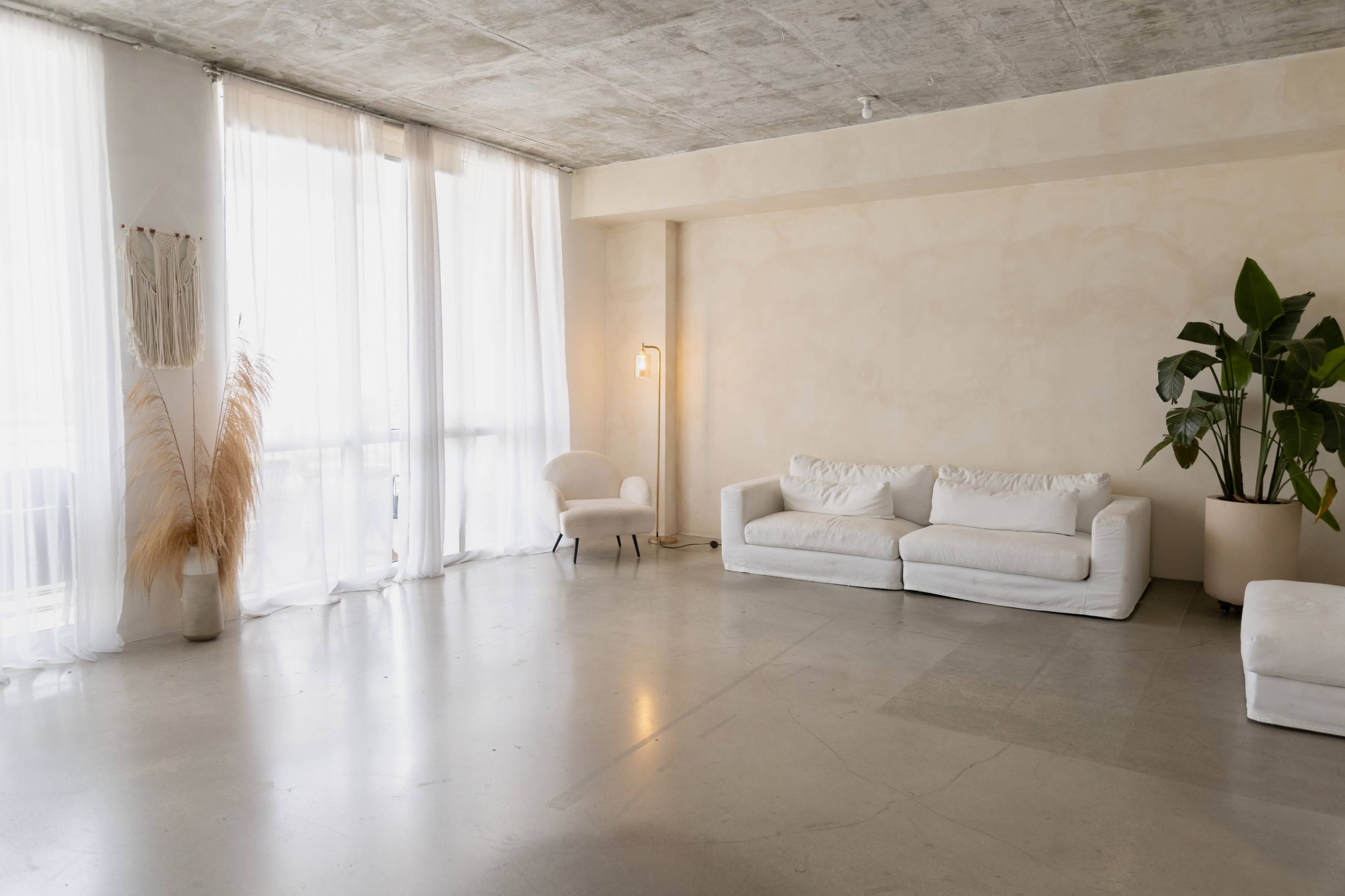 A spacious, minimally furnished room features a white sofa, a single armchair, tall curtains, and a large potted plant.