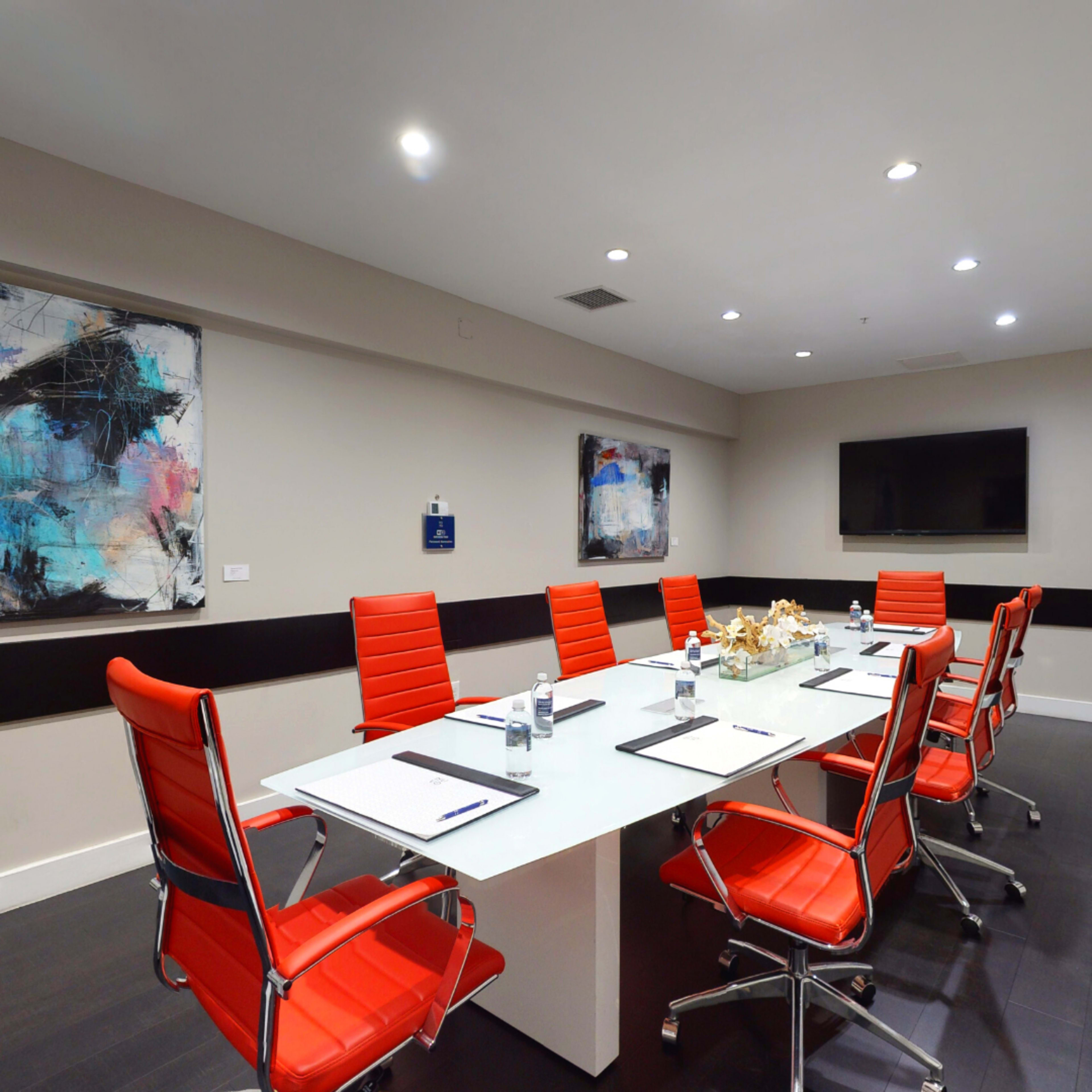 A conference room features a large white table surrounded by red chairs, with artwork on the walls and a television at one end.