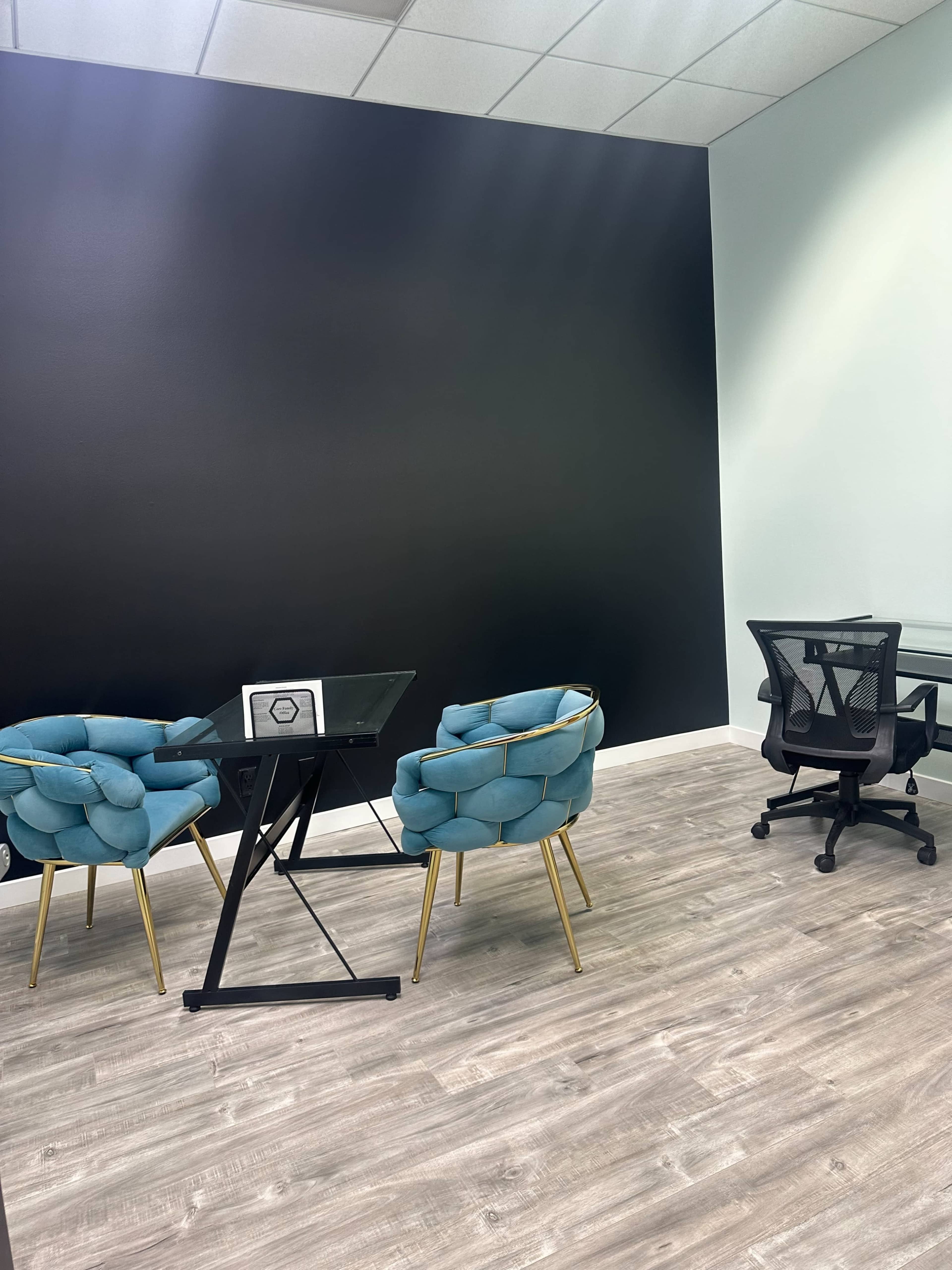 A small office space features two upholstered chairs, a glass table, and a black accent wall.