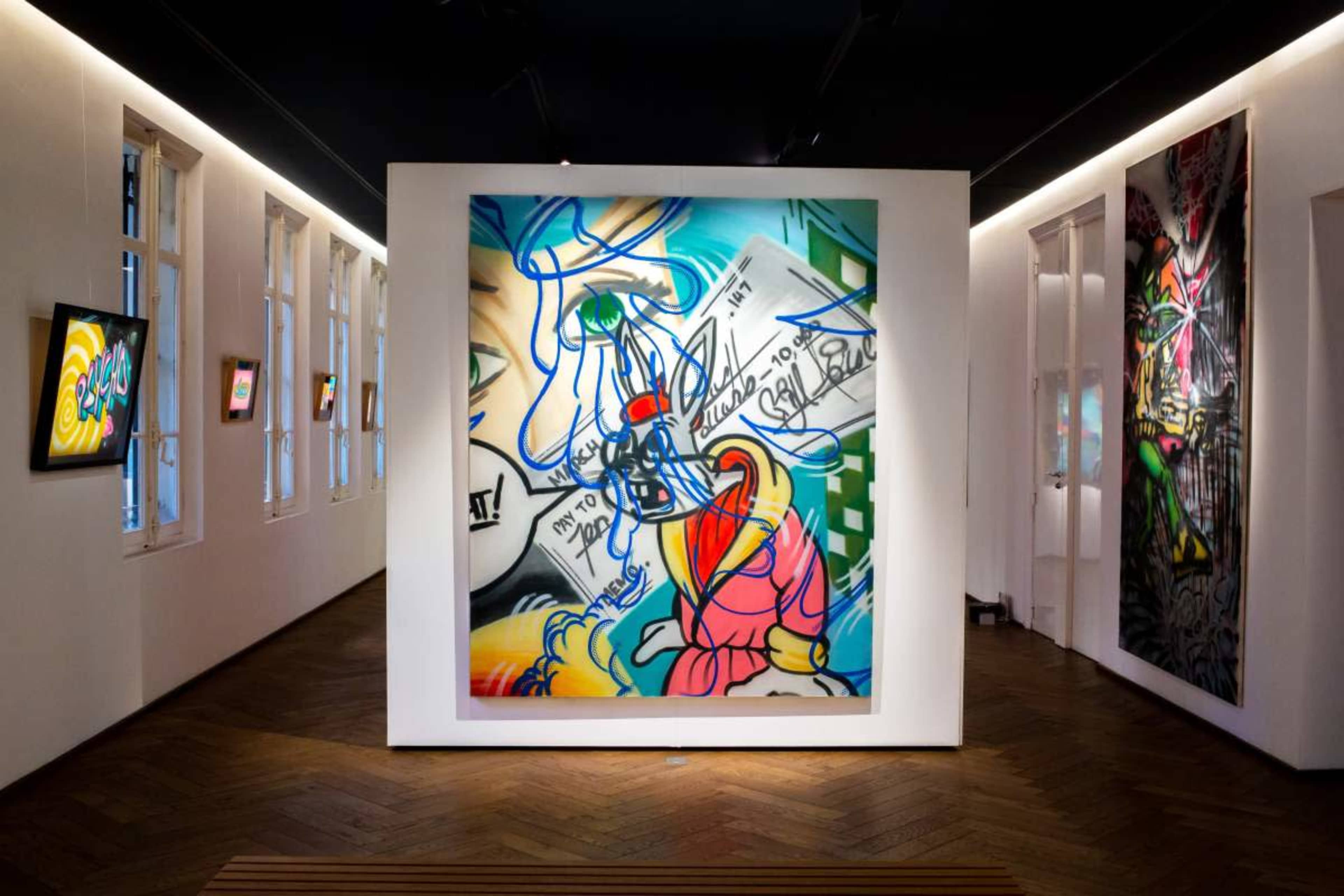 The image shows an art gallery featuring a large colorful painting on a central wall, with smaller framed artworks displayed on adjacent walls.
