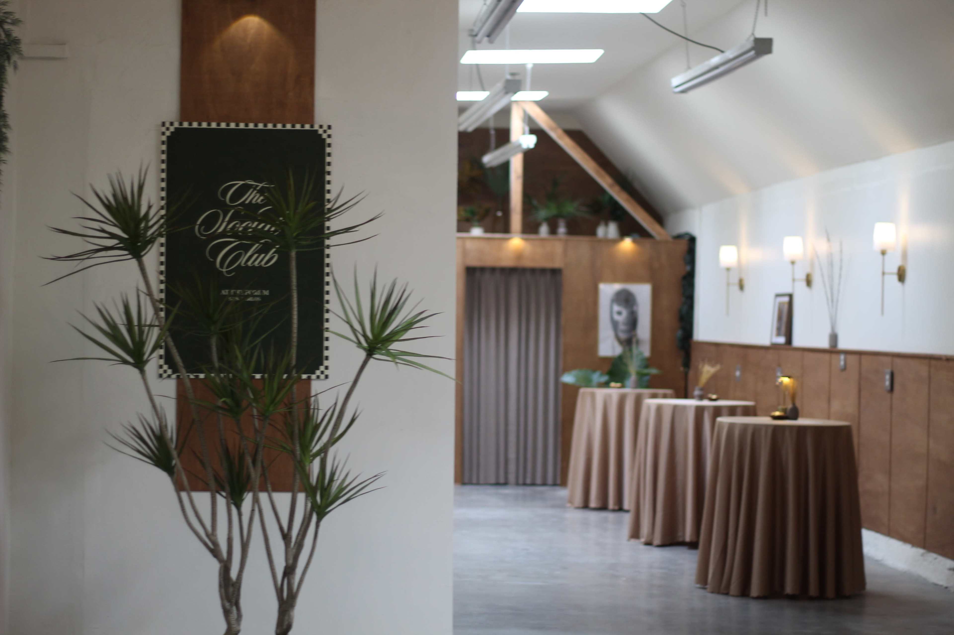 The image shows an indoor event space featuring a sign for "The Secret Club," tables with beige tablecloths, and a decorative plant in the foreground.