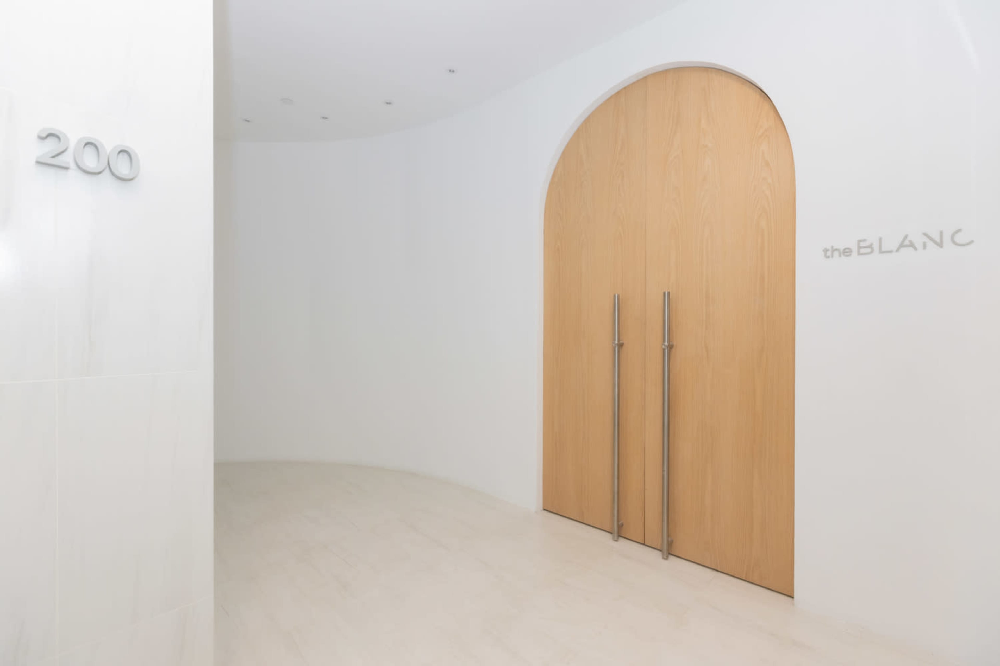 The image shows a modern entrance to a room with wooden double doors and a curved wall adorned with the number "200" and the name "the BLANC."