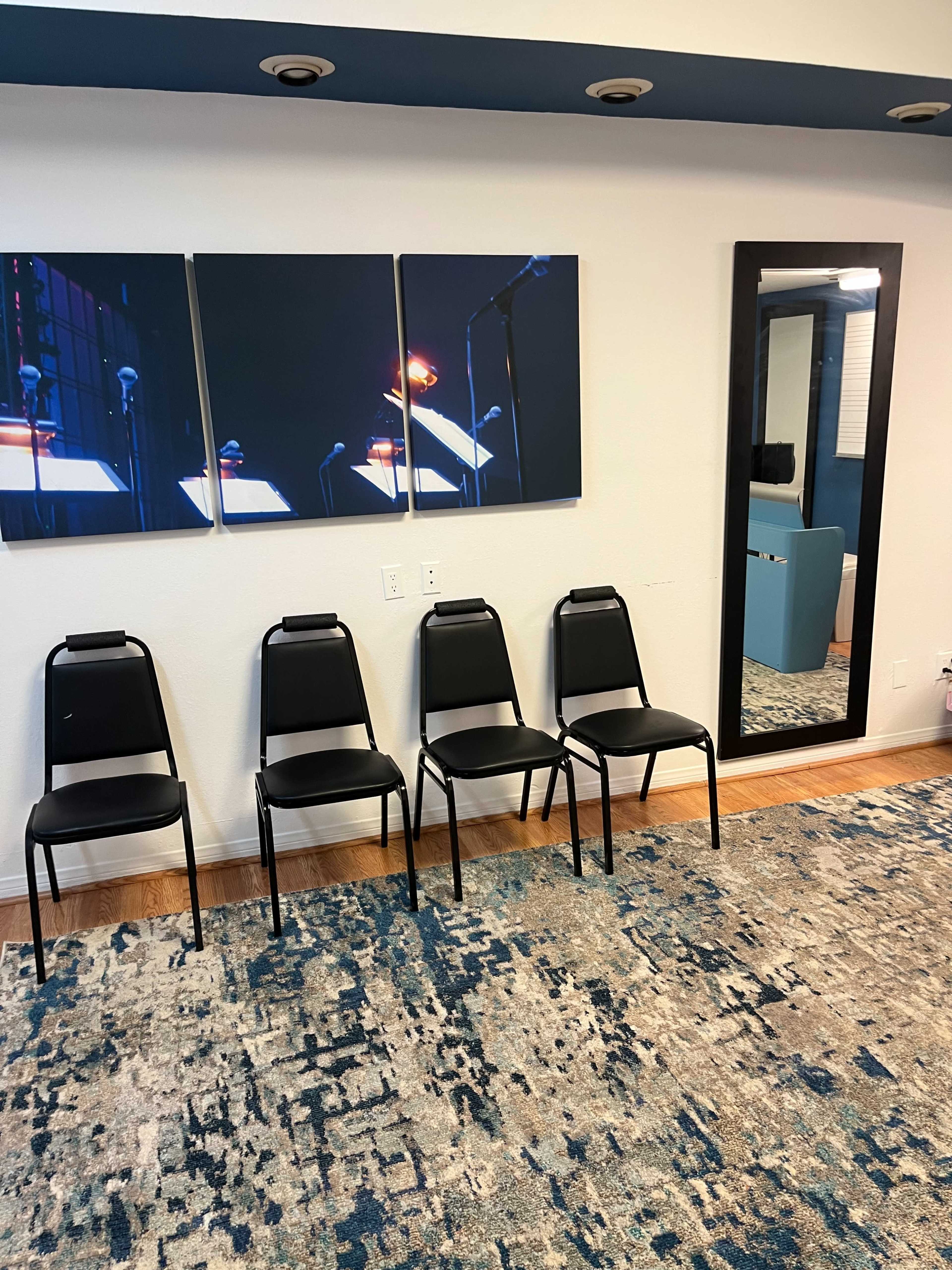 A row of four black chairs lined up against a wall with three framed pictures above them and a large mirror adjacent to the chairs.