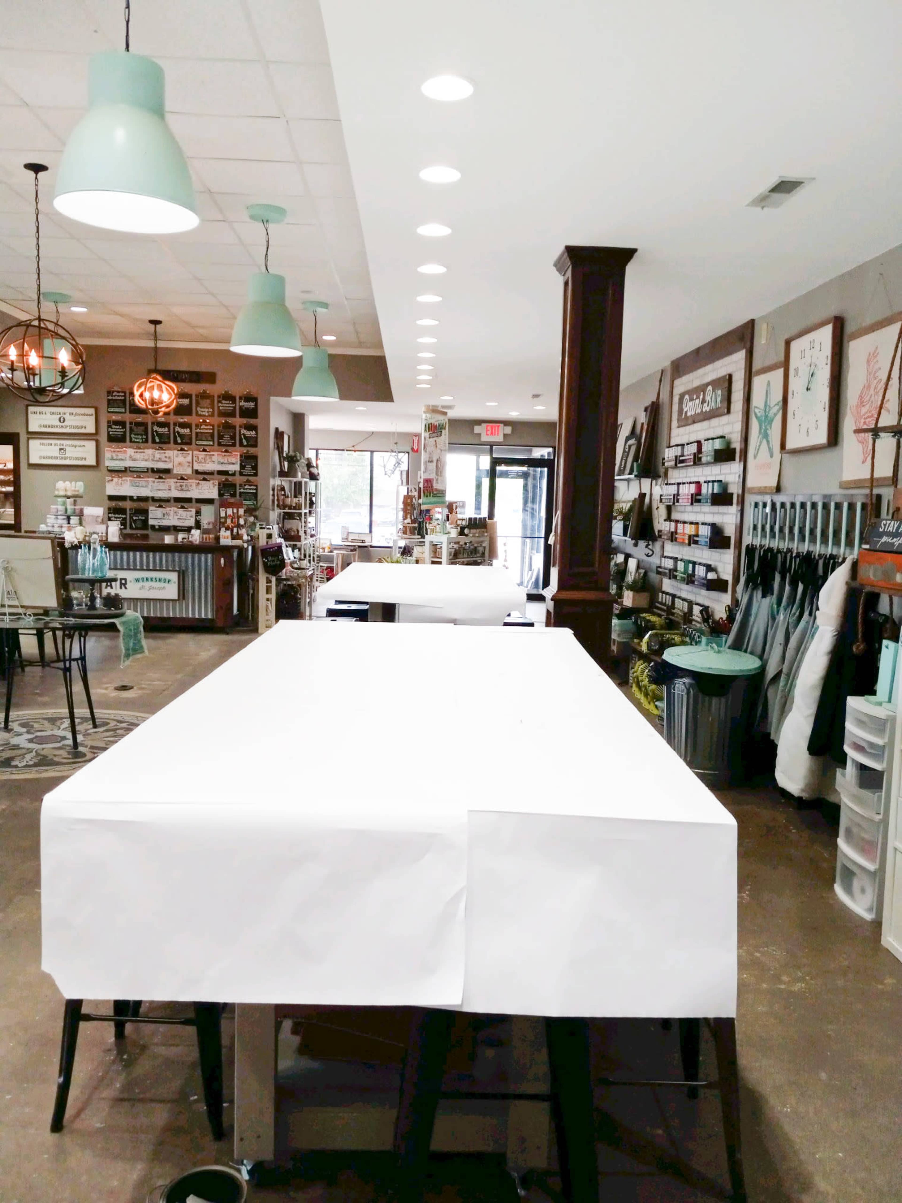 A crafting store interior with several empty tables covered in white tablecloths, lined by shelves displaying various art supplies and decorations.
