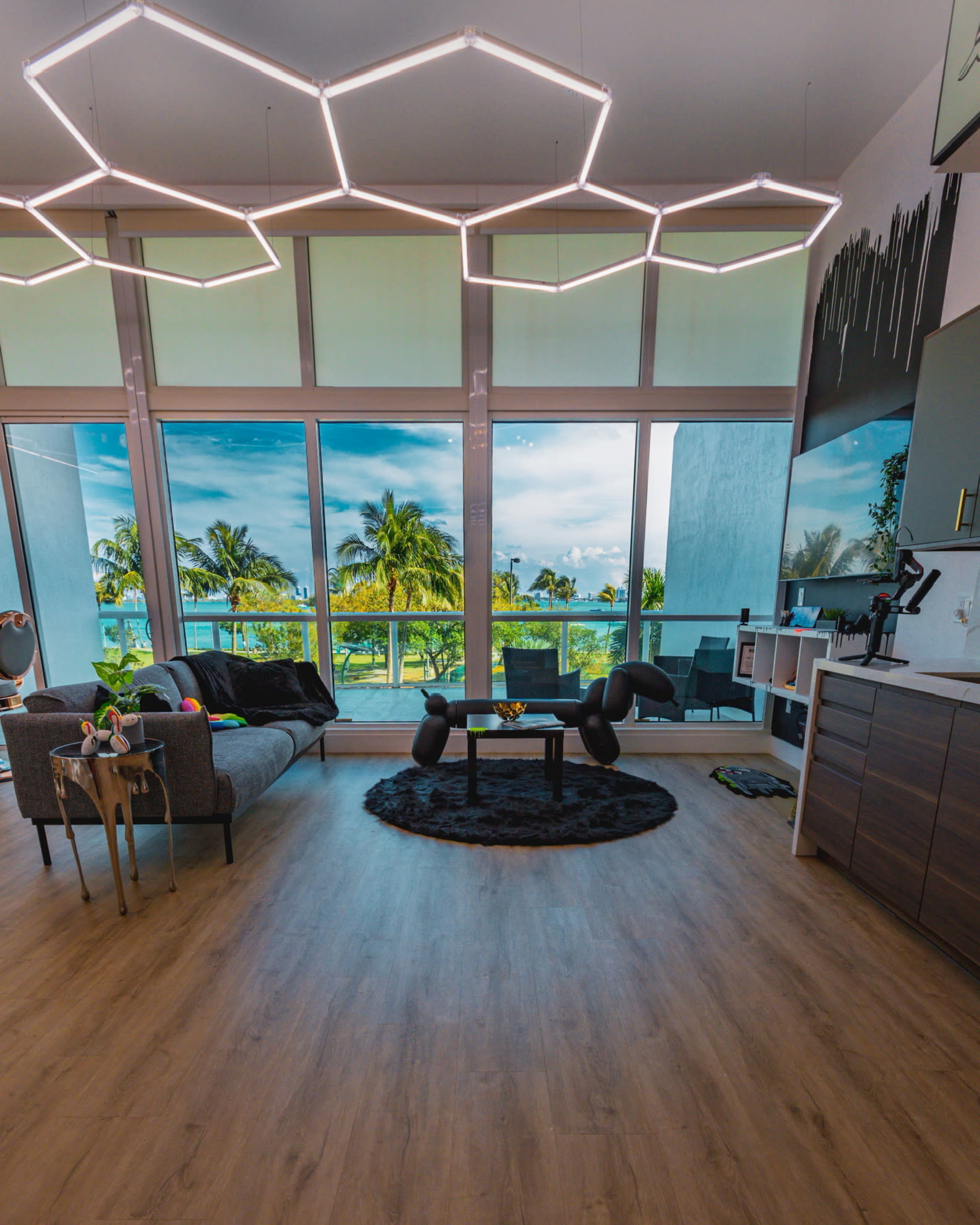 A modern living room features a sofa, a coffee table on a rug, and large windows showcasing palm trees and a blue sky outside.