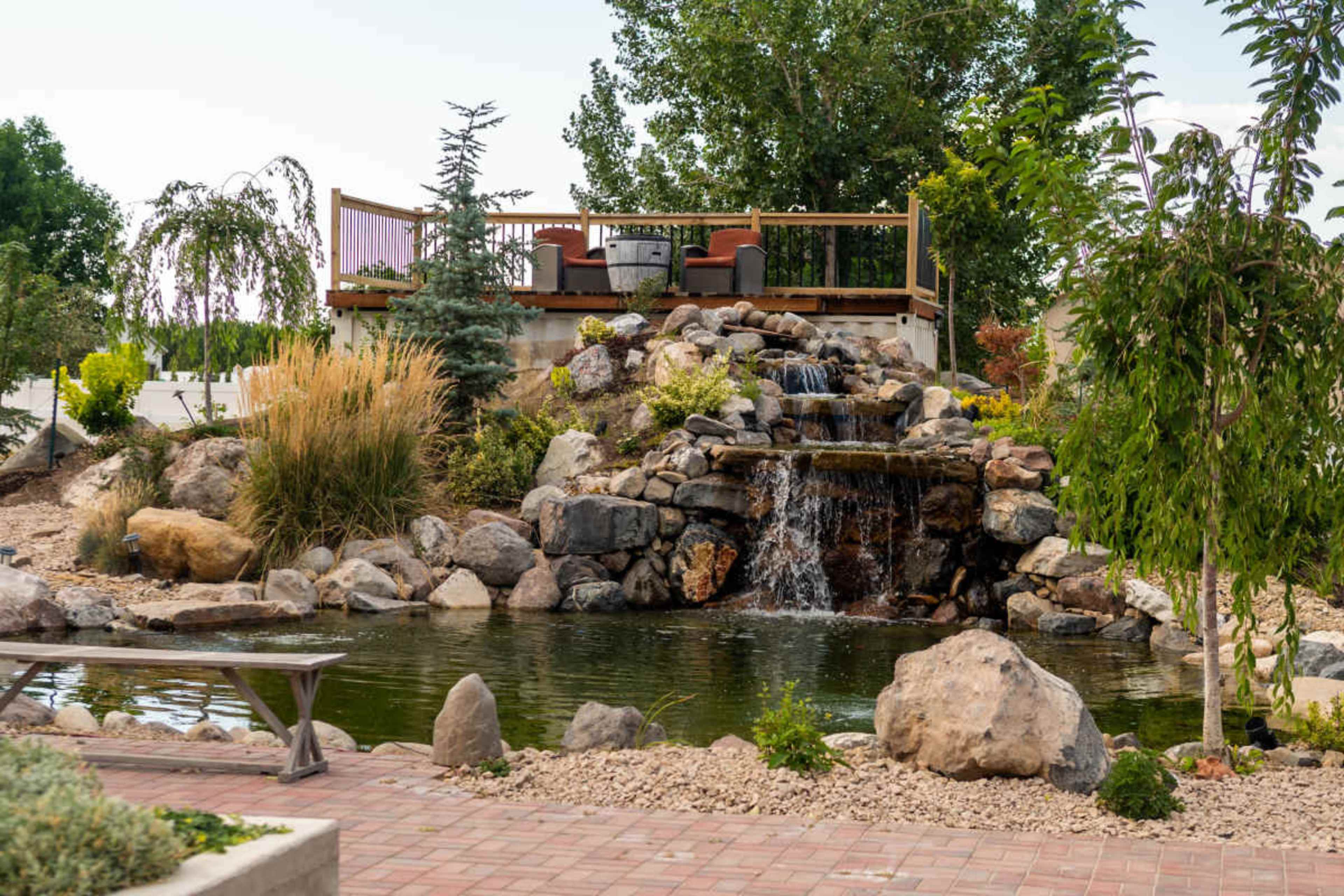 The image shows a landscaped garden featuring a rock waterfall cascading into a pond, with a deck and seating area in the background surrounded by various plants and trees.