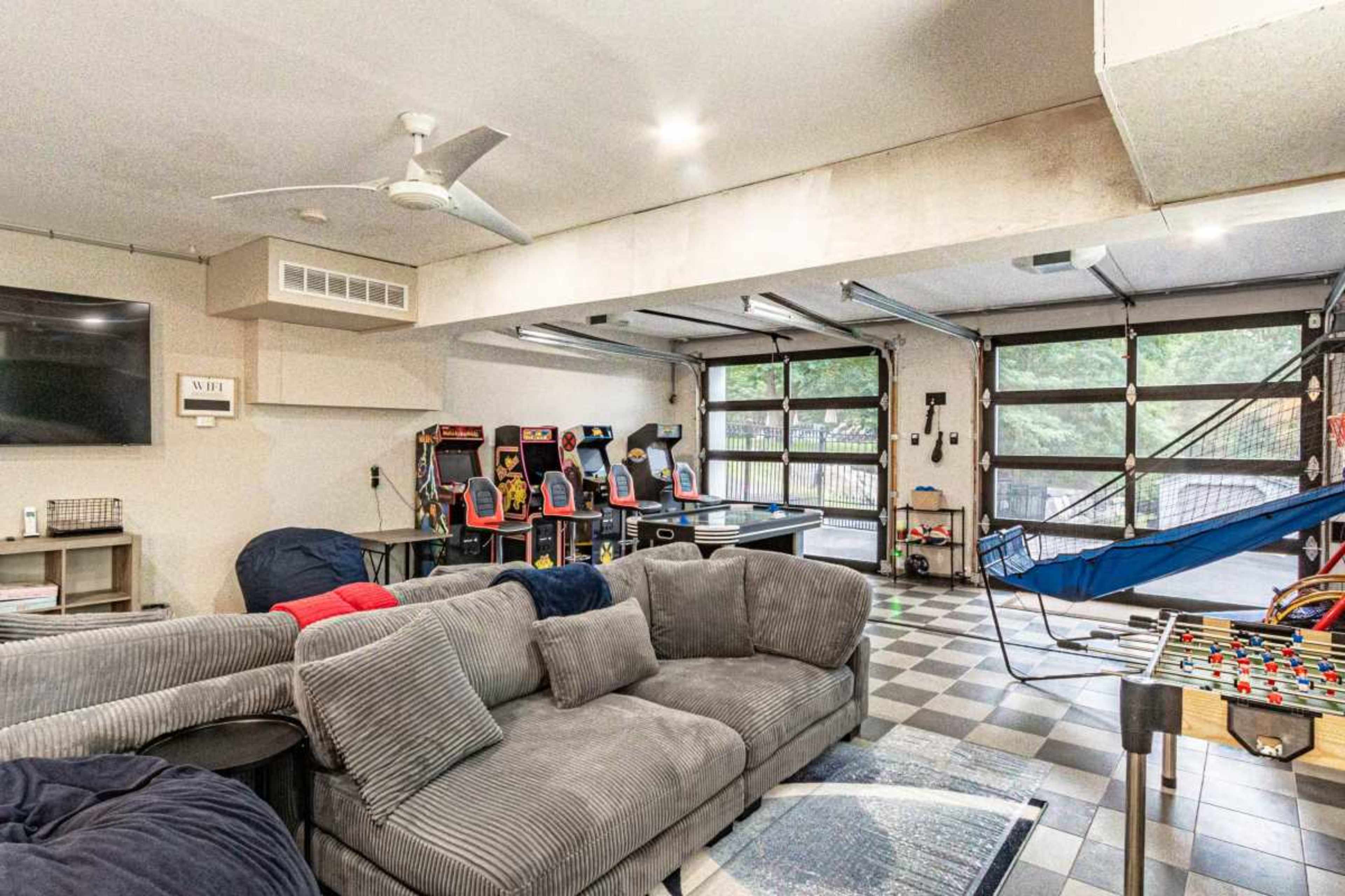 The image depicts a spacious game room featuring arcade machines, a foosball table, a ping pong table, a large sectional couch, and a suspended hammock.