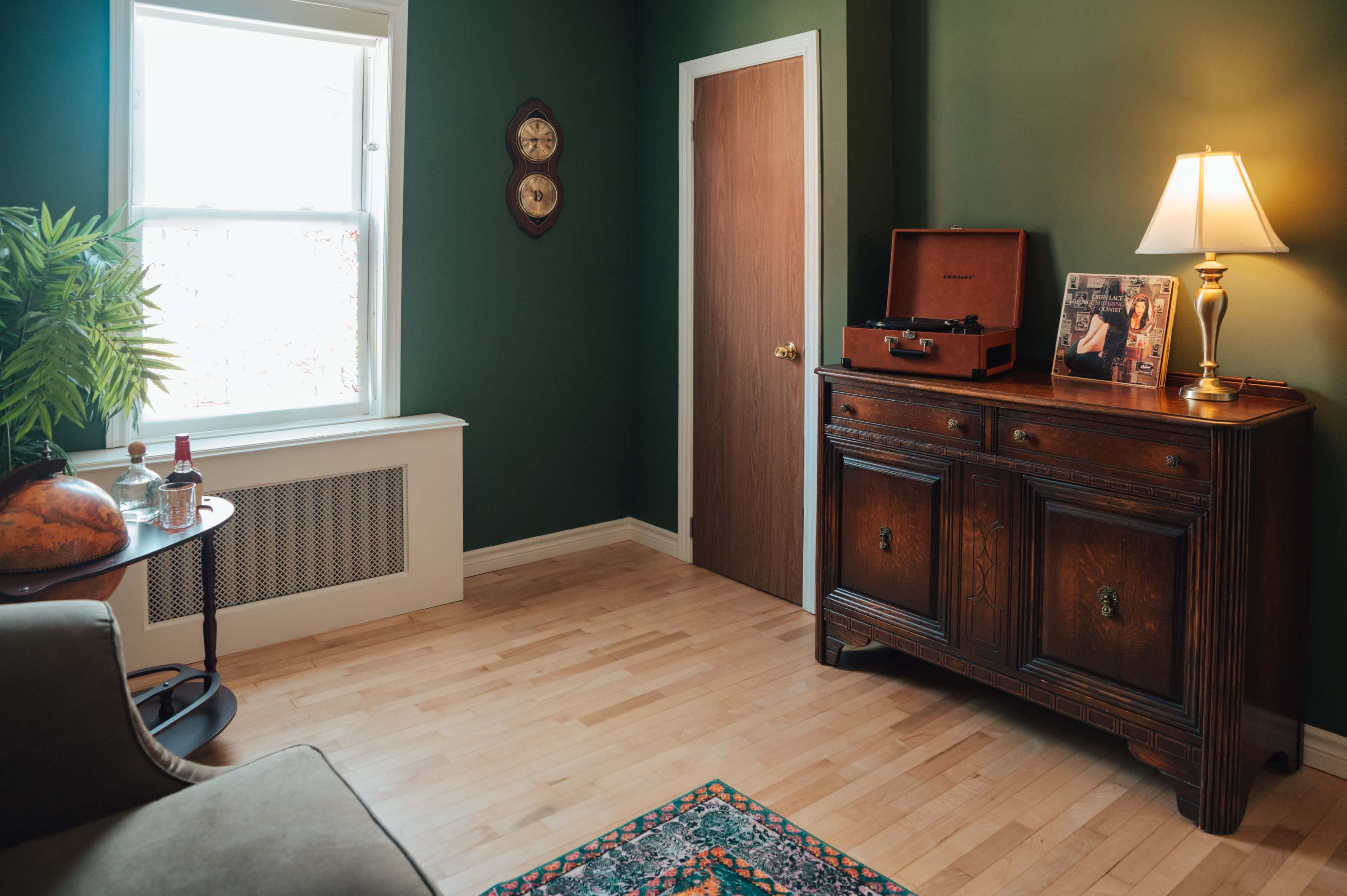 The image shows a cozy room with green walls, featuring a wooden cabinet, a record player, a lamp, and a window letting in natural light.