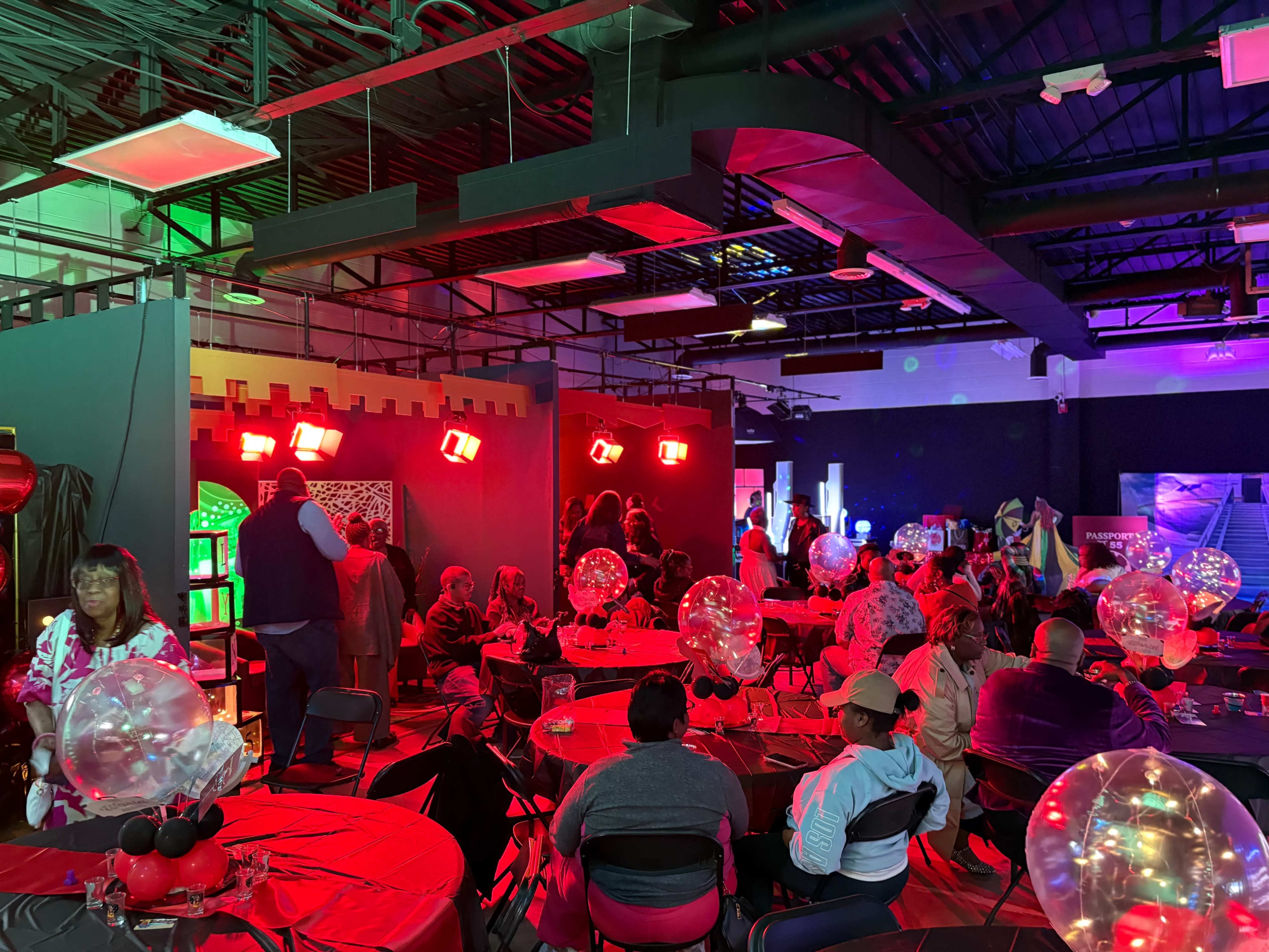 A diverse group of people gathers in a brightly lit event space adorned with colorful decorations and tables set with balloons.