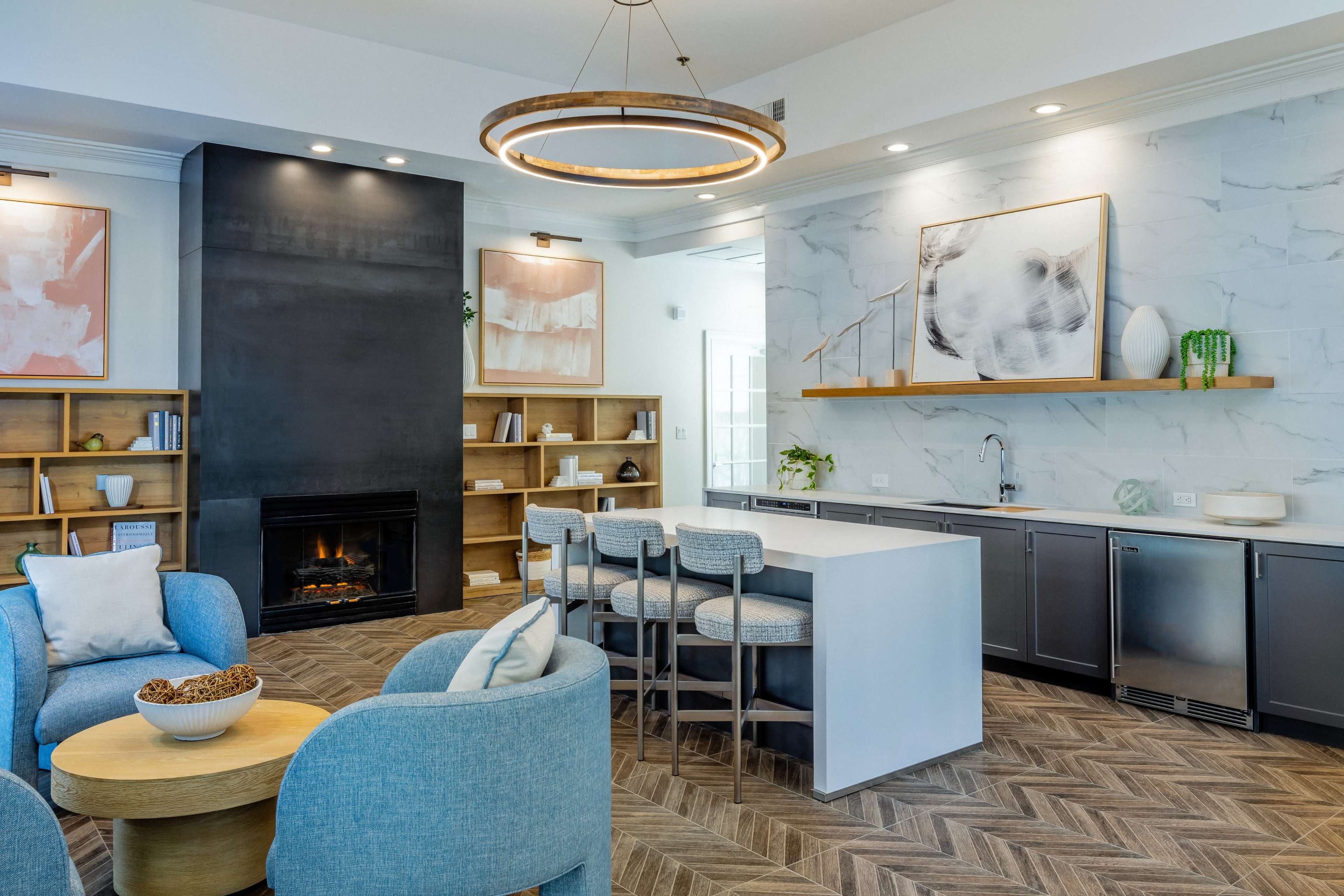 A modern living space featuring a sleek kitchen with gray cabinetry, a large island with seating, a fireplace, and a cozy seating area with light blue chairs.
