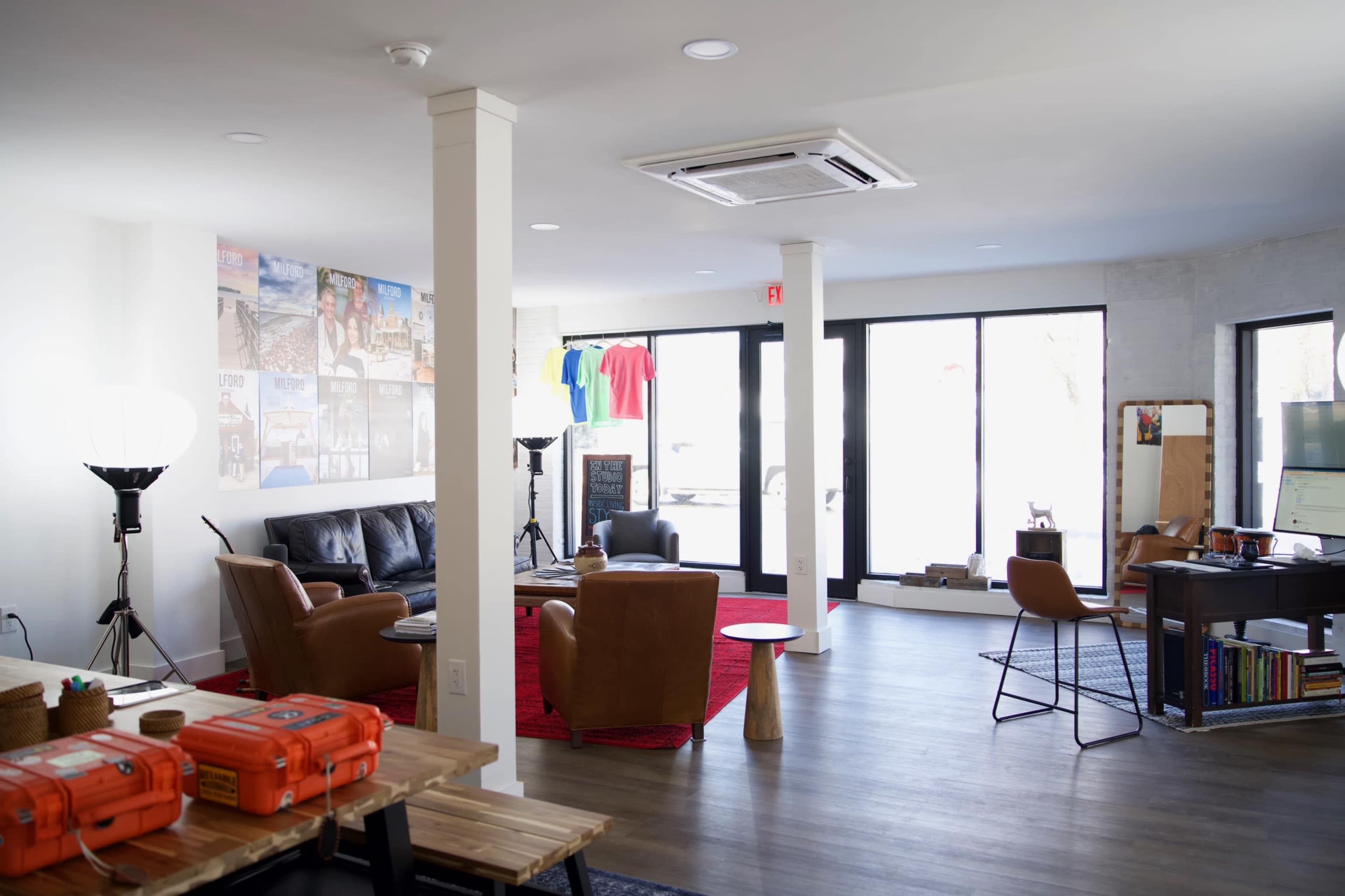 The image shows a modern, open-concept office space featuring a lounge area with leather chairs, a small table, and a desk, along with large windows allowing natural light.