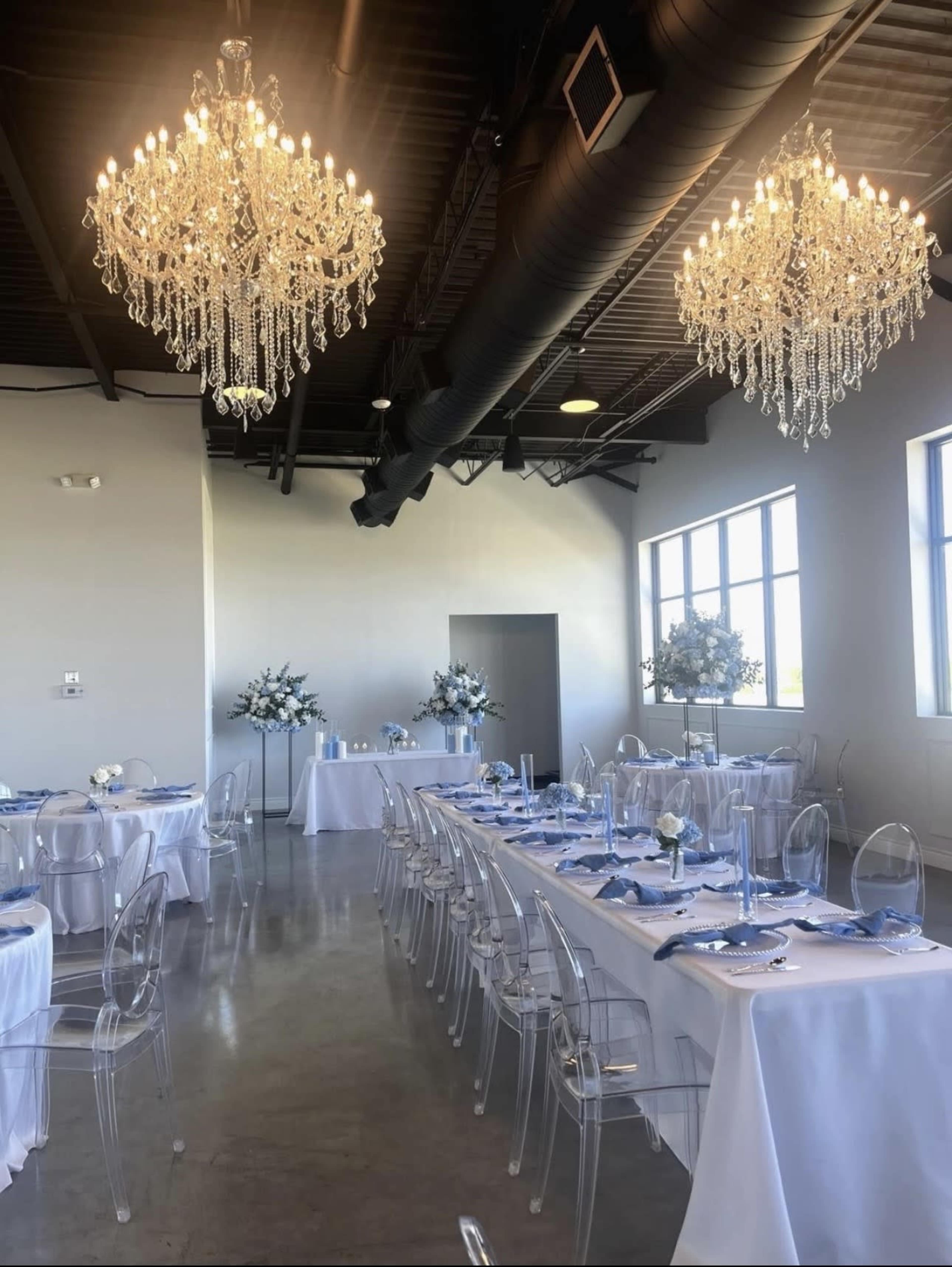 A well-appointed dining area features clear acrylic chairs, white tablecloths, and elegant crystal chandeliers, with a floral centerpiece on each table.