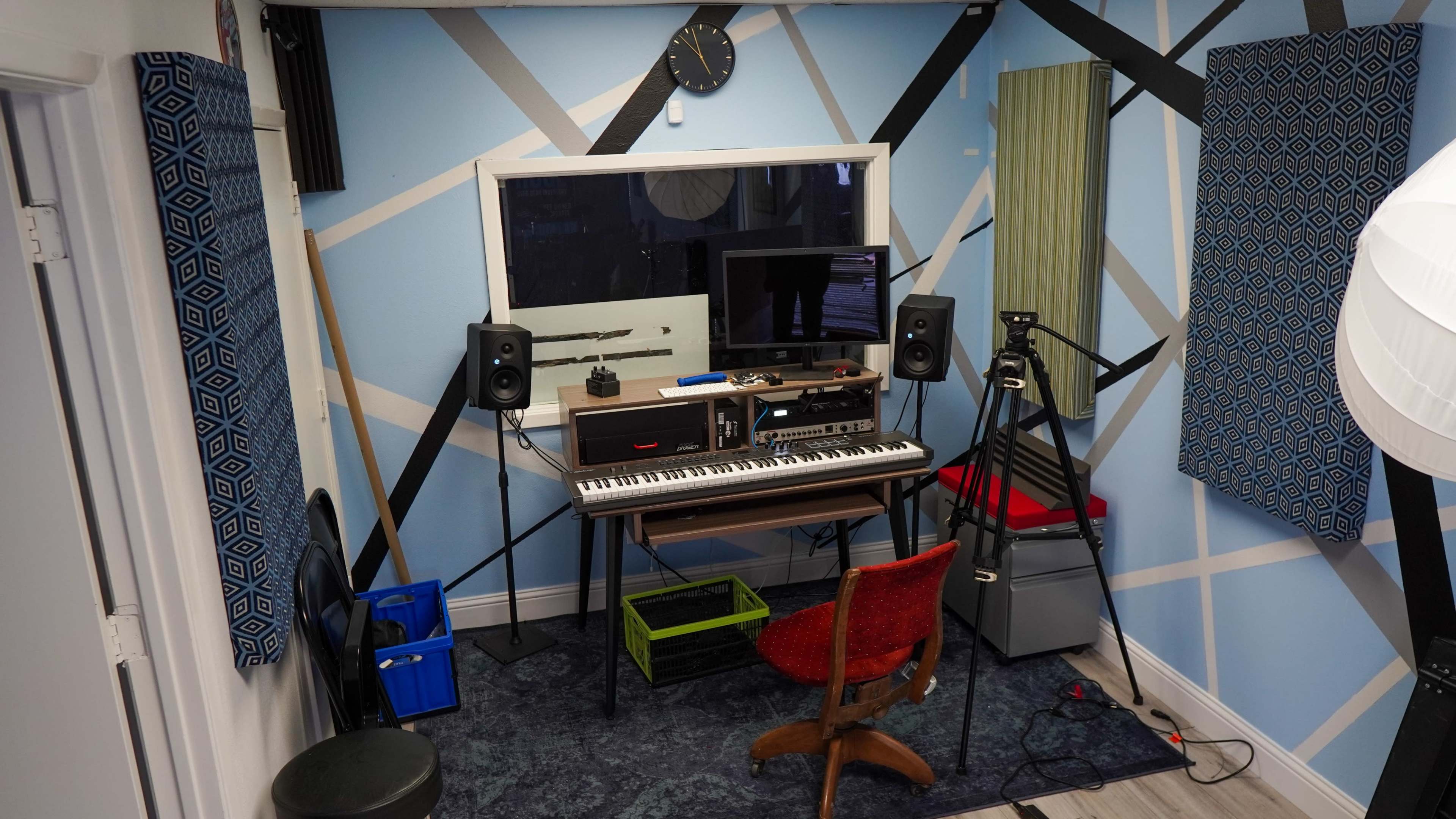 A music recording studio with a keyboard, computer monitor, speakers, and a chair, set against a patterned blue wall.