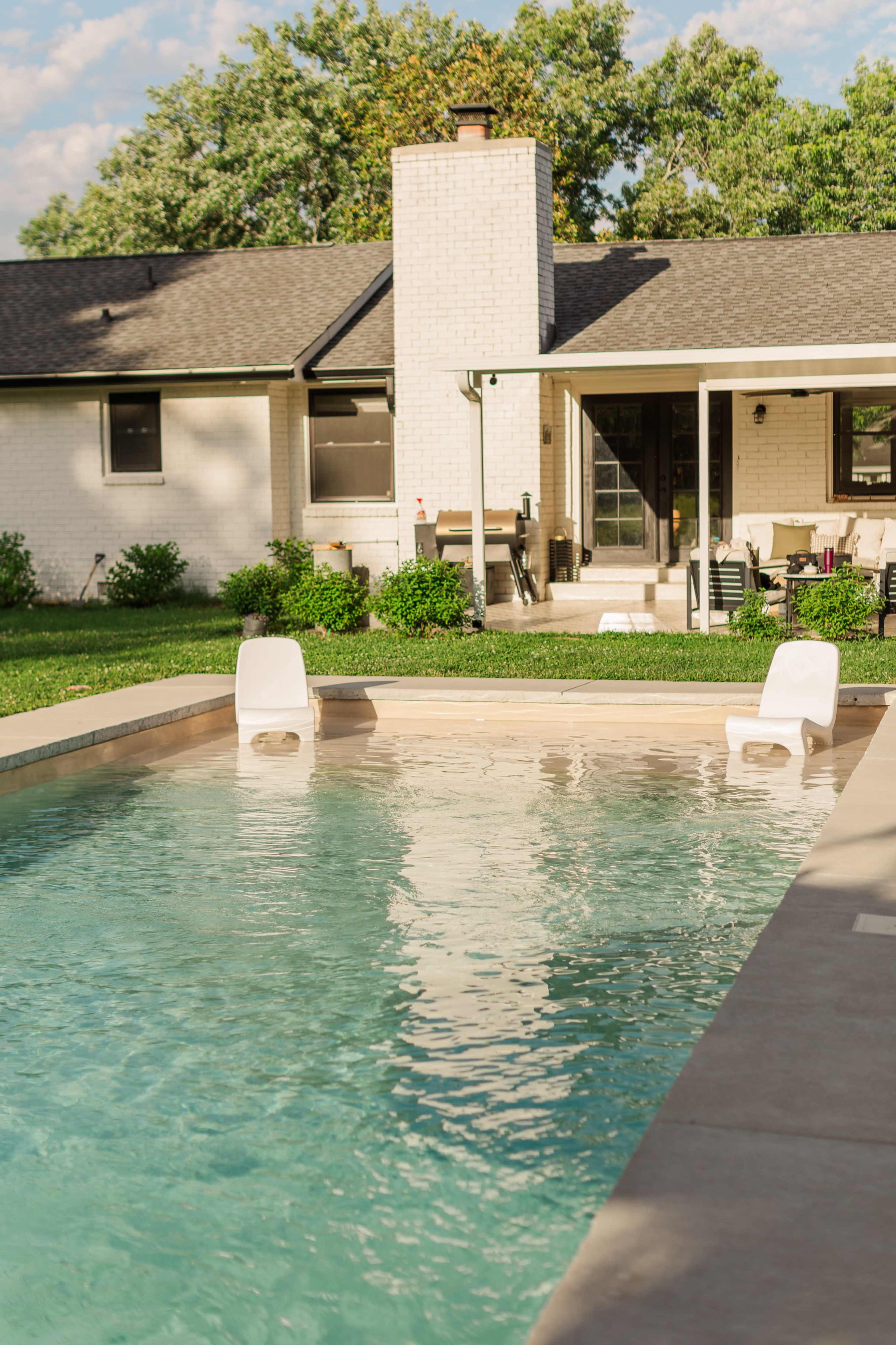 The image shows a backyard swimming pool with two white lounge chairs, a house in the background, and a patio area covered by an awning.