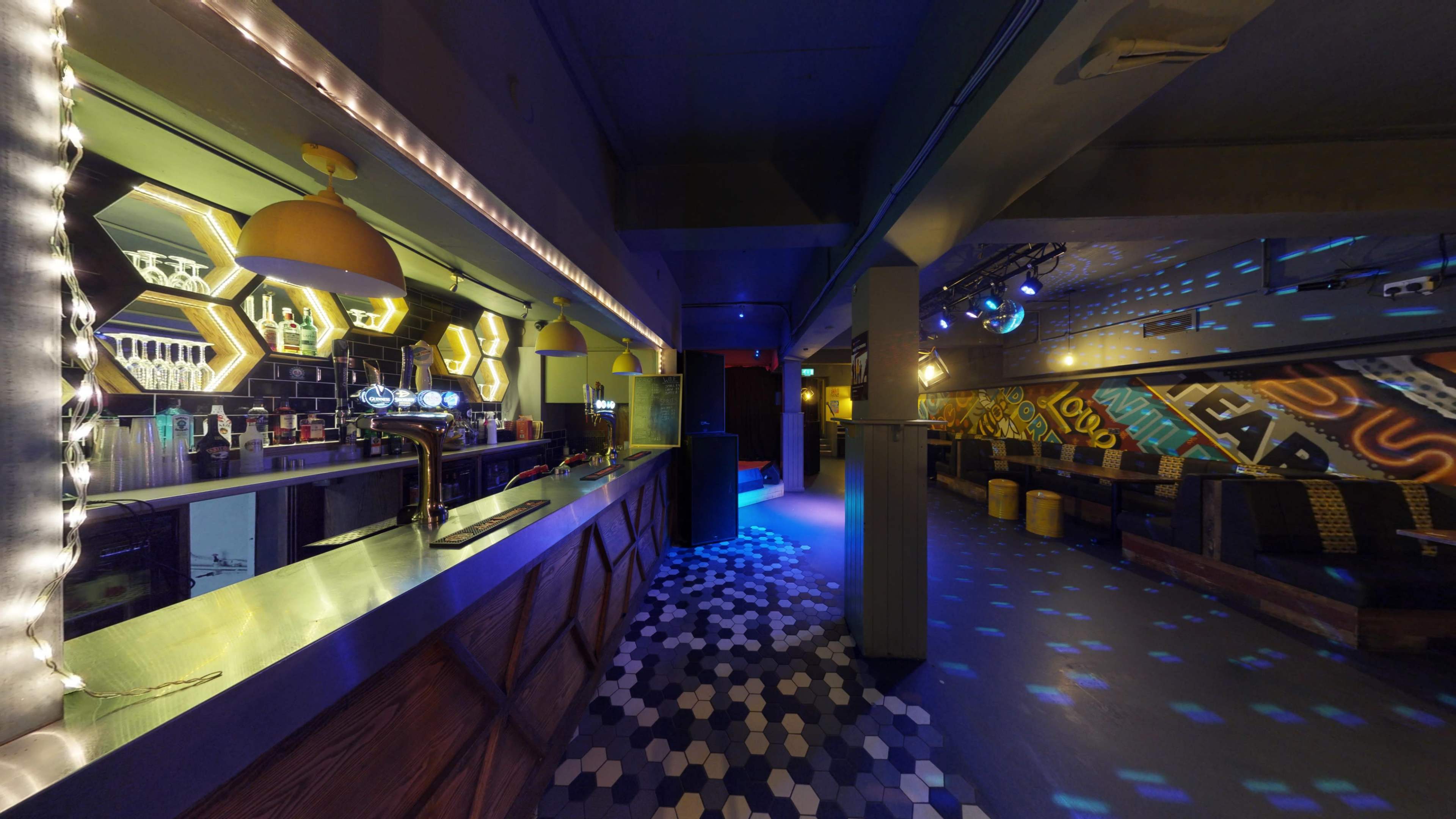 An interior view of a bar with a long counter, colorful lighting, and a patterned floor.