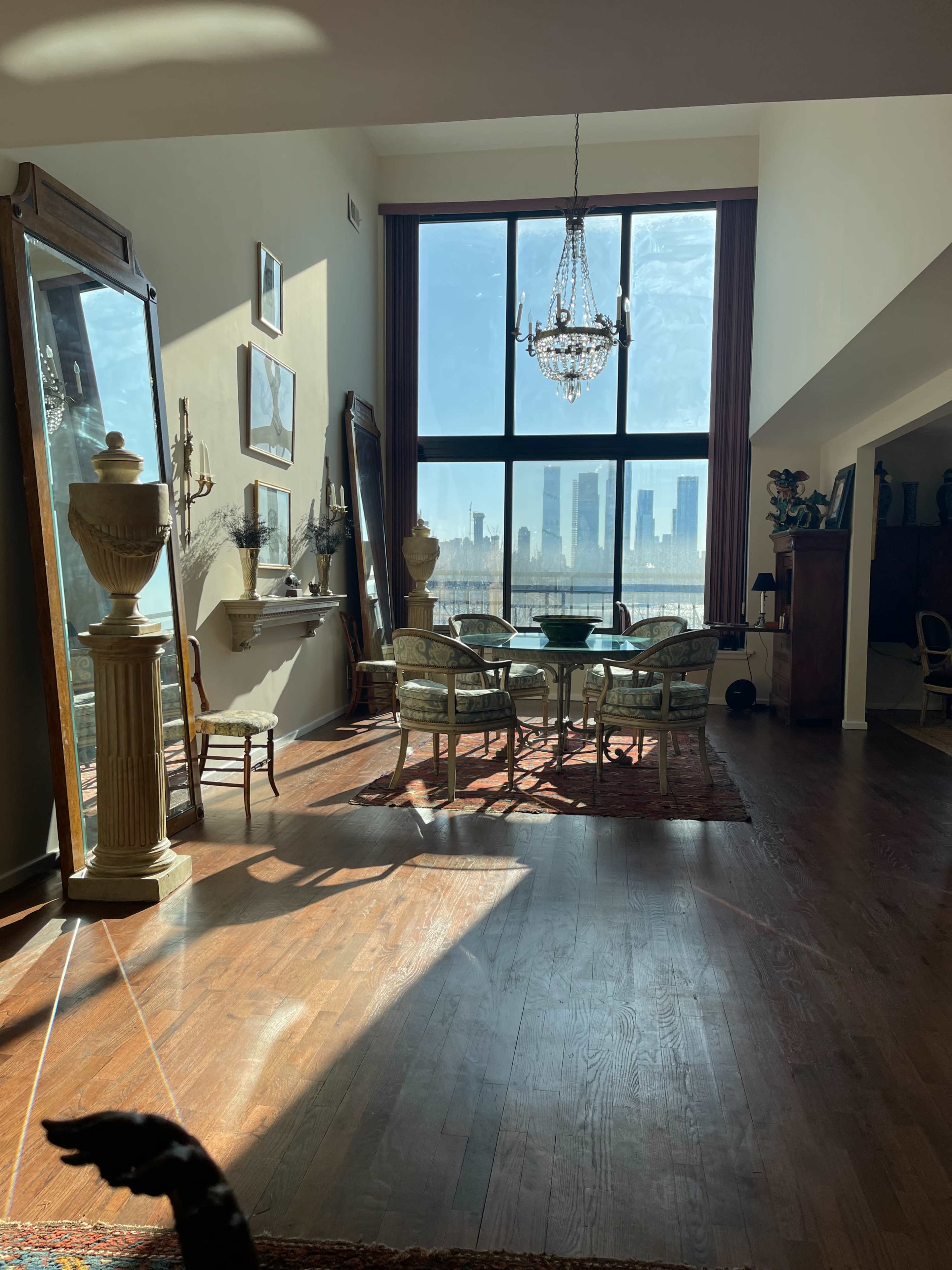 A sunlight-filled room features a dining table surrounded by chairs, a large mirror, and a view of city skyscrapers through expansive windows.