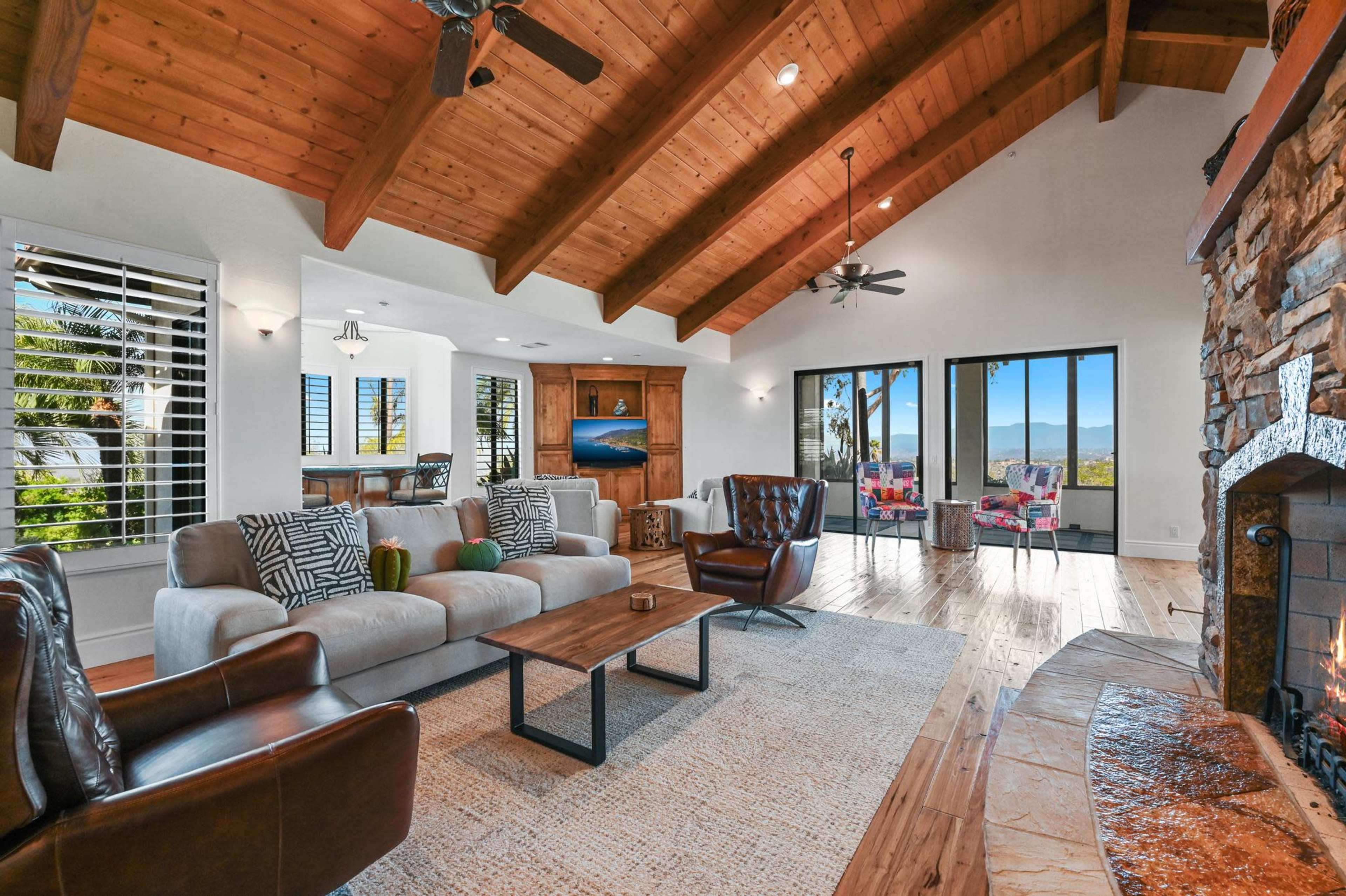 A spacious living room with exposed wooden beams, a stone fireplace, and large windows opening to a view of the outdoors.