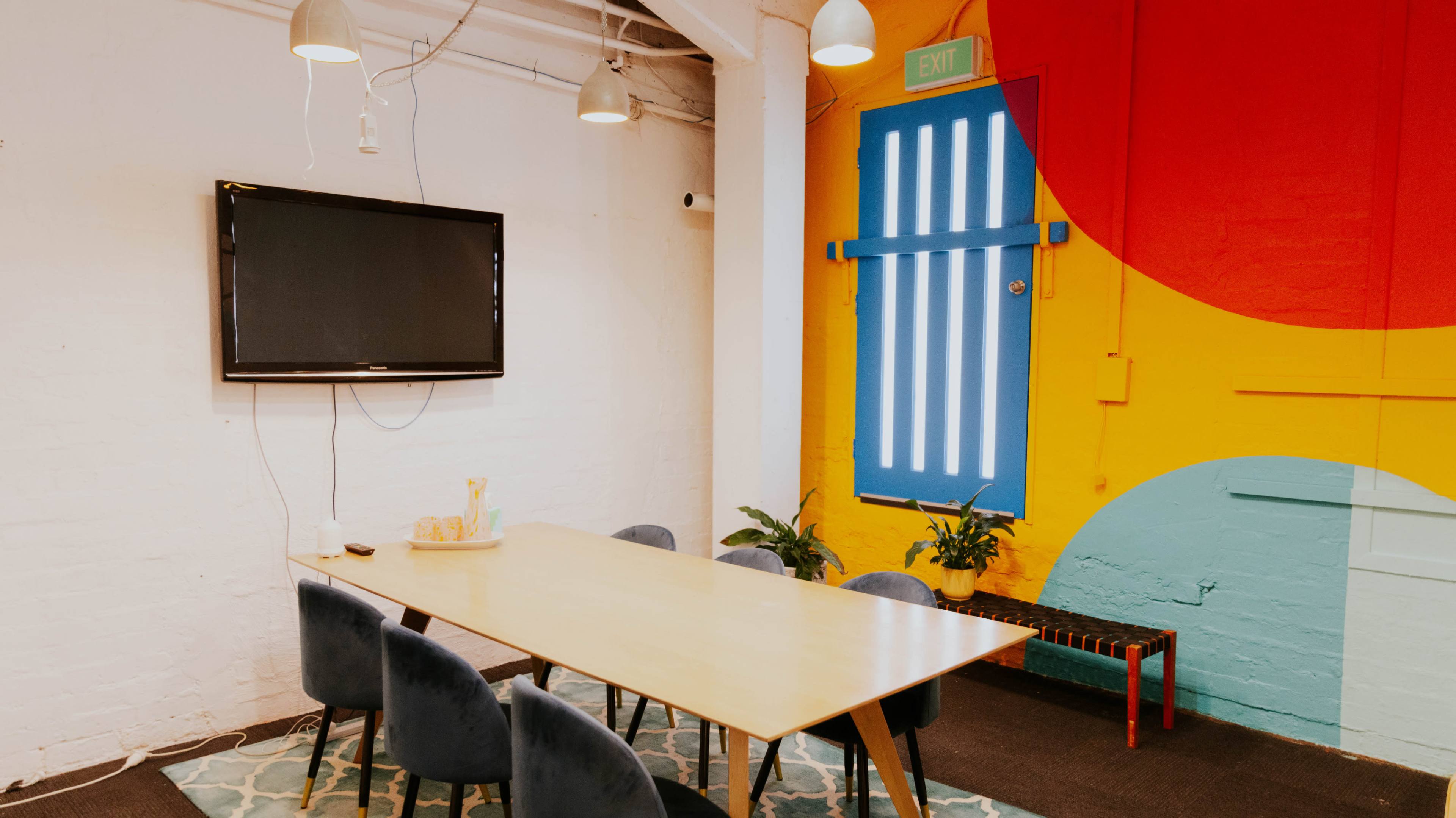 A bright meeting room features a large wooden table surrounded by six blue chairs, a wall-mounted television, and a colorful mural with a blue door and vibrant design.