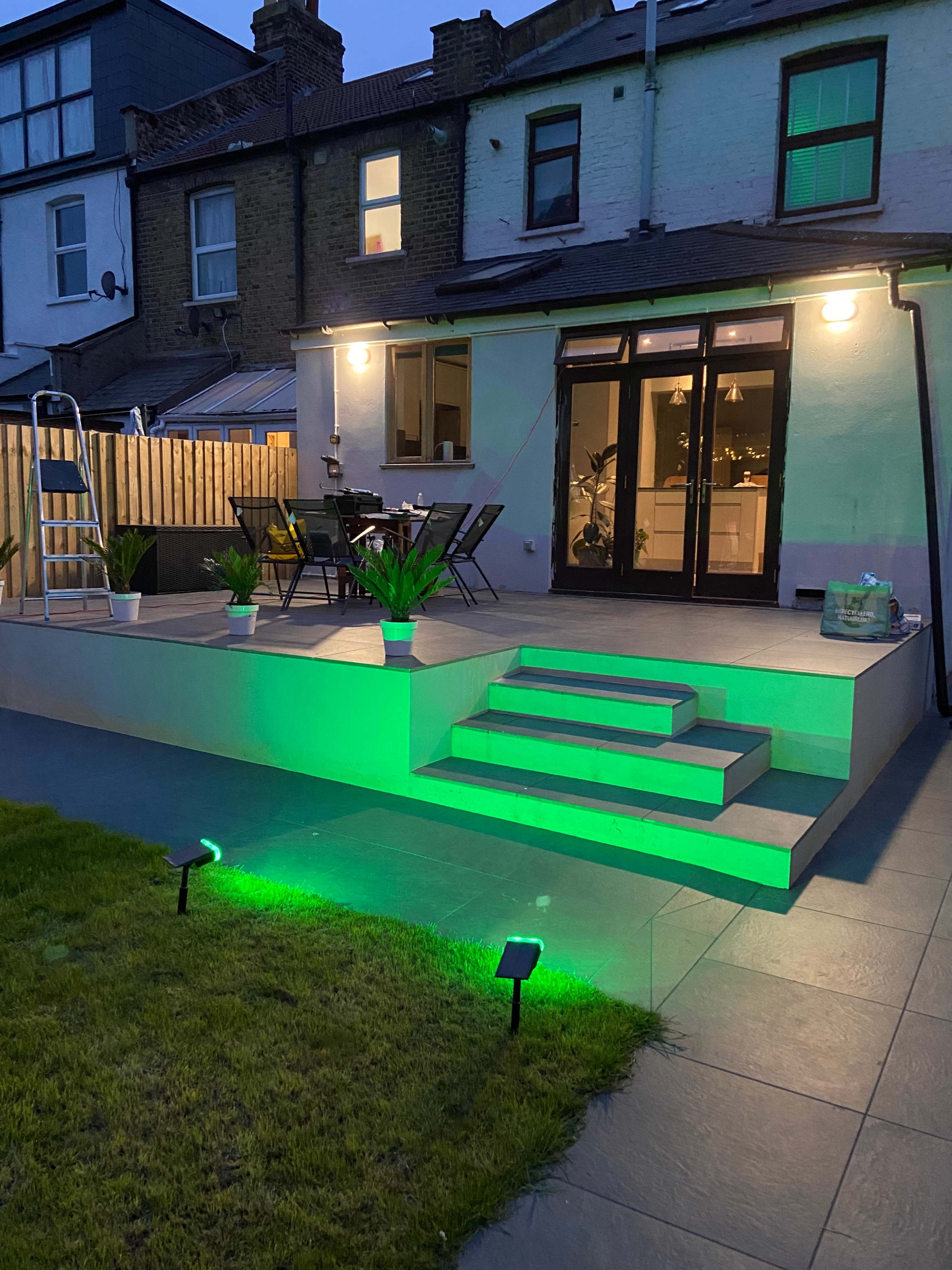A backyard patio features green lighting, steps leading to the entrance, and seating arrangements near the house.