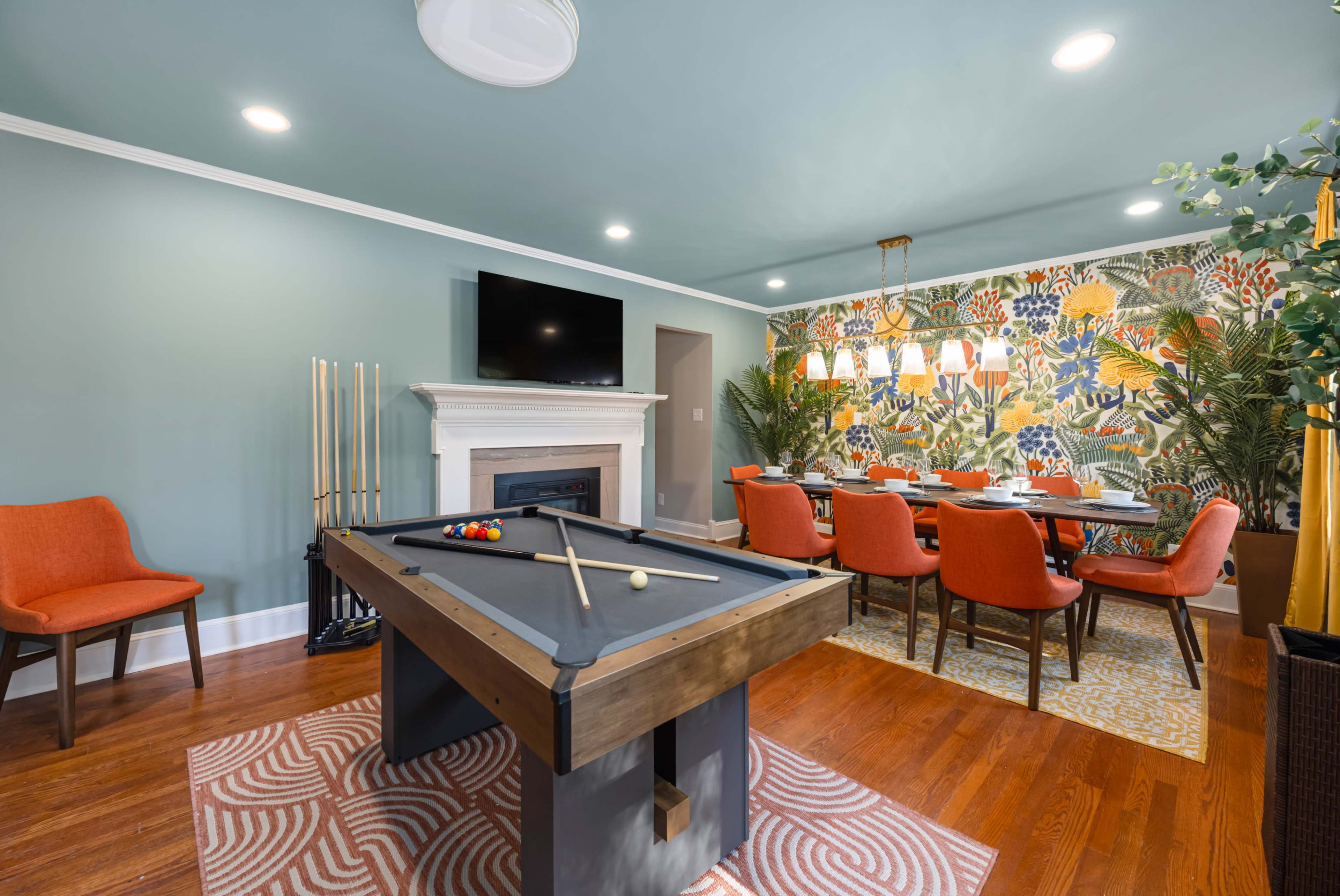 The image shows a modern game room featuring a pool table in the foreground, a fireplace, a large dining table with chairs, and a vibrant floral wallpaper in the background.