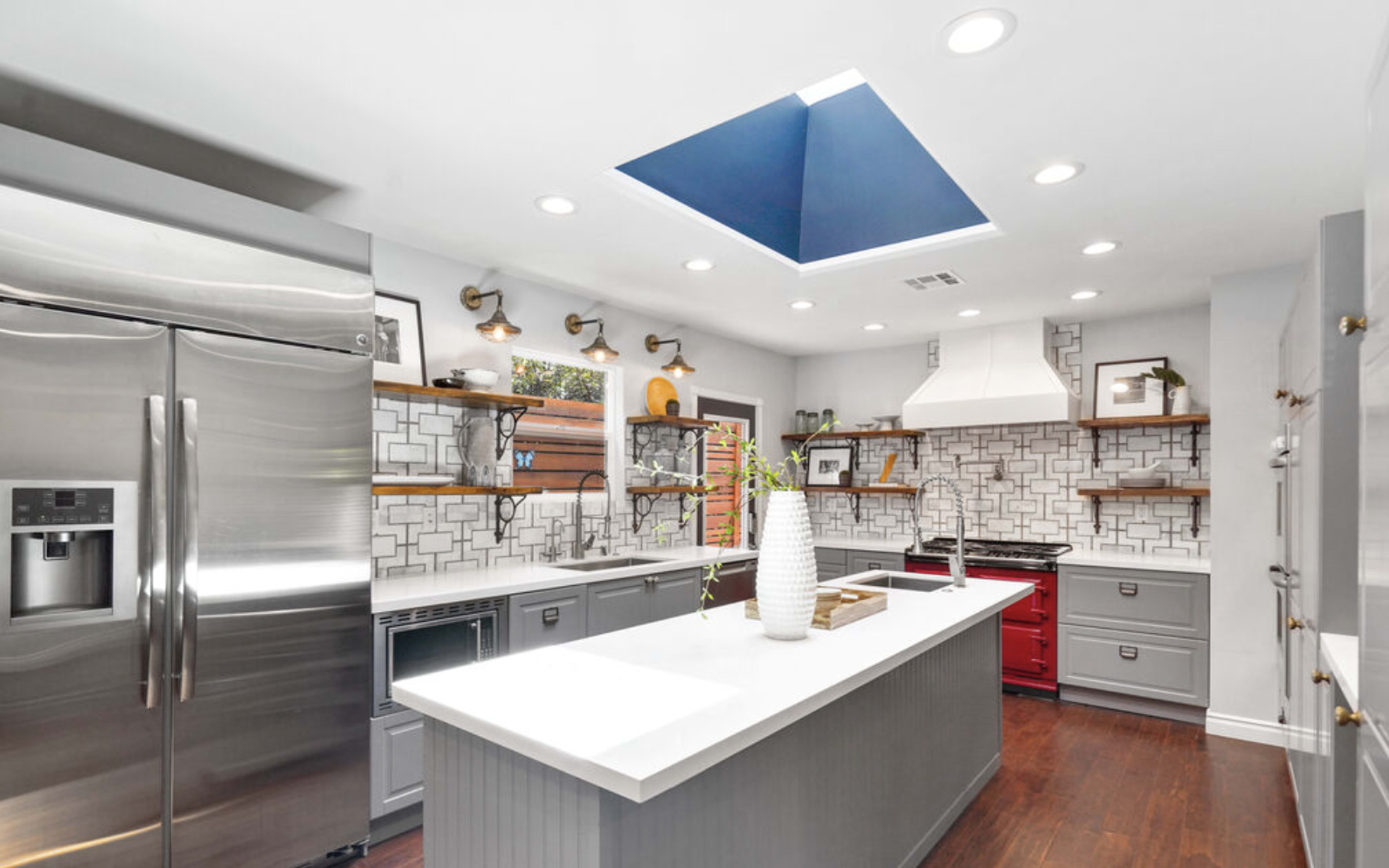 A modern kitchen features a center island with a white countertop, stainless steel appliances, and a blue skylight overhead.