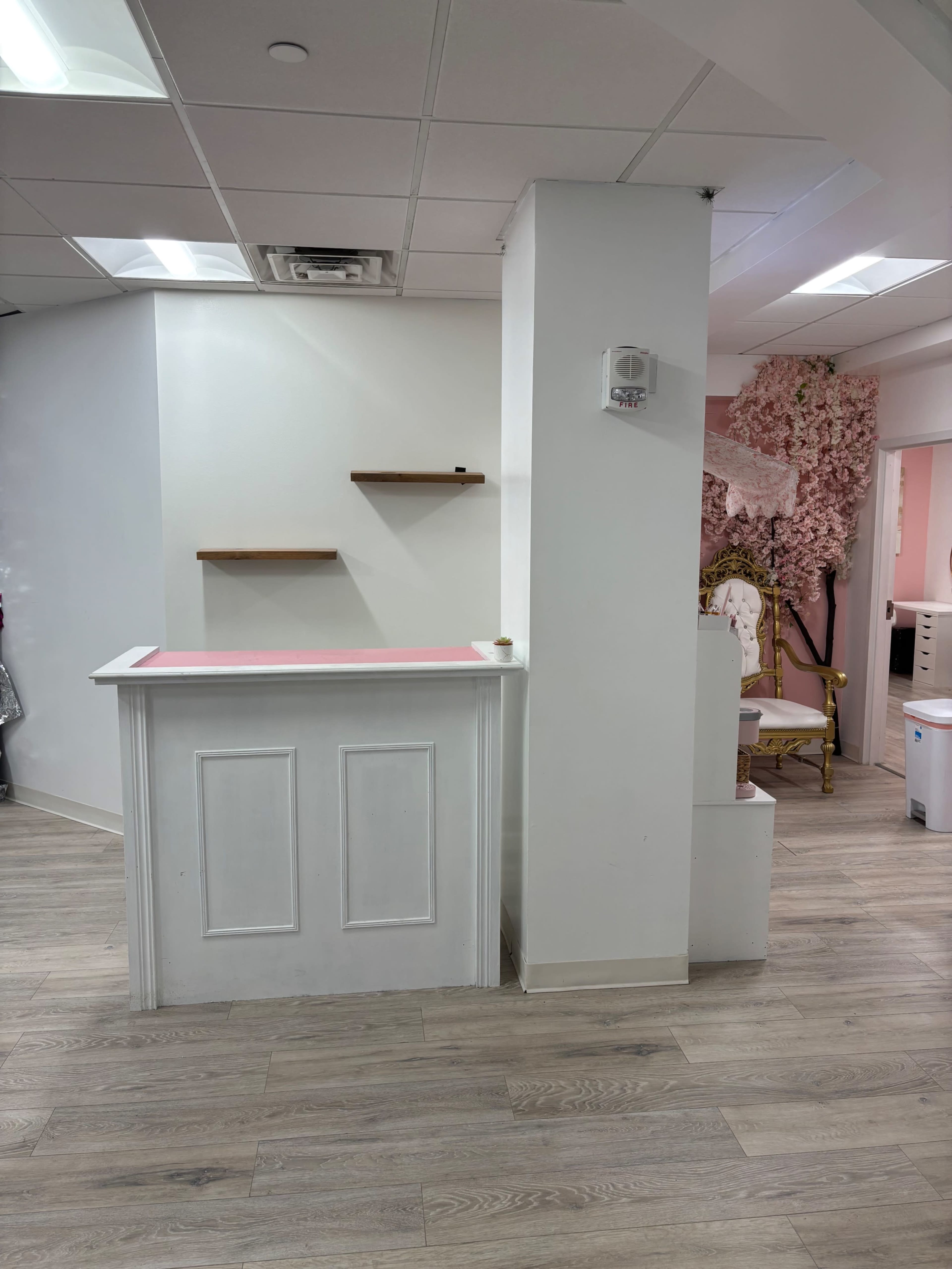 The image shows an interior of a reception area featuring a white front desk, wooden shelves on the wall, and pink accents in the decor.