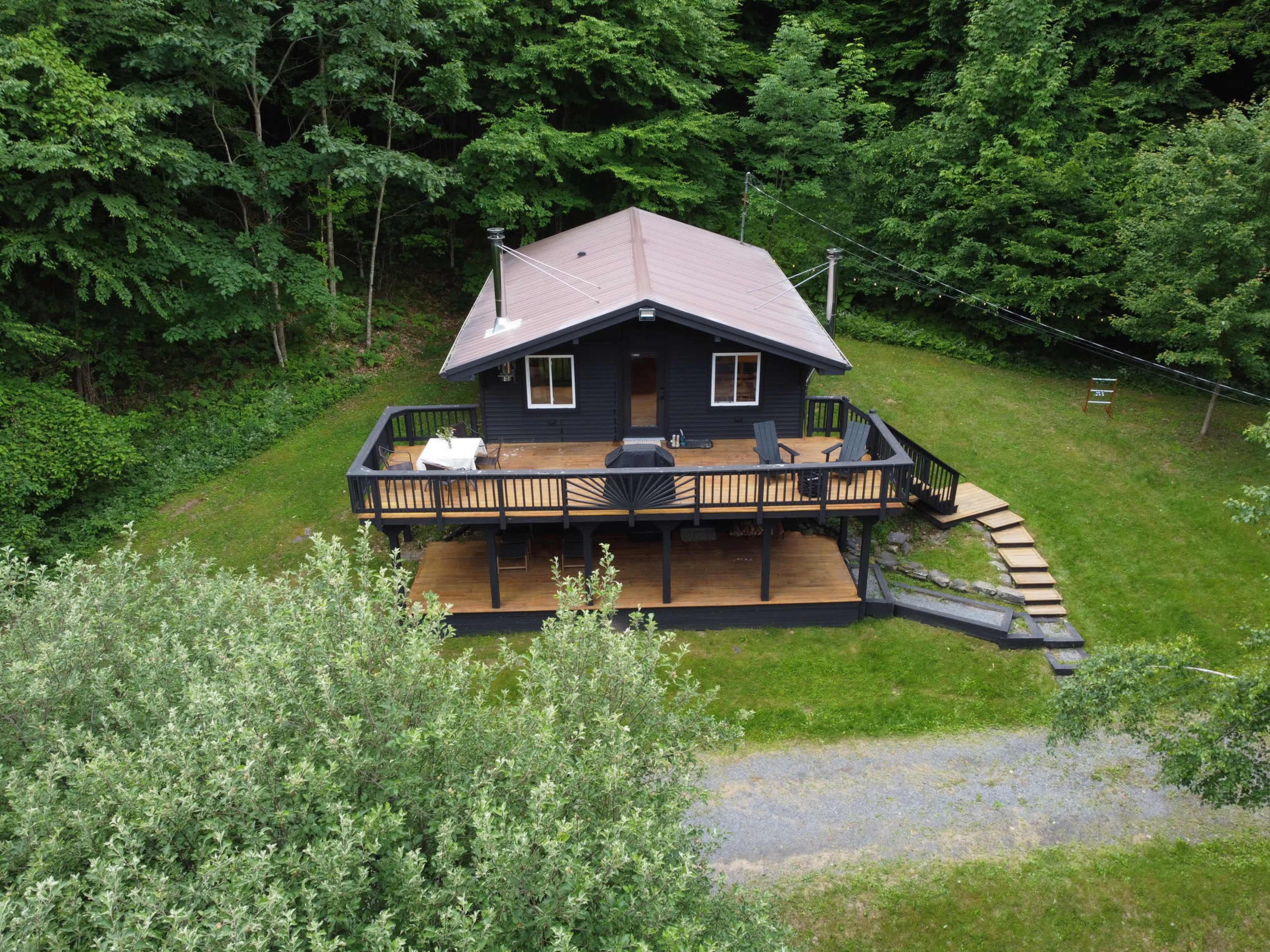 A modern black cabin with a wooden deck is surrounded by greenery and has a pathway leading to it.