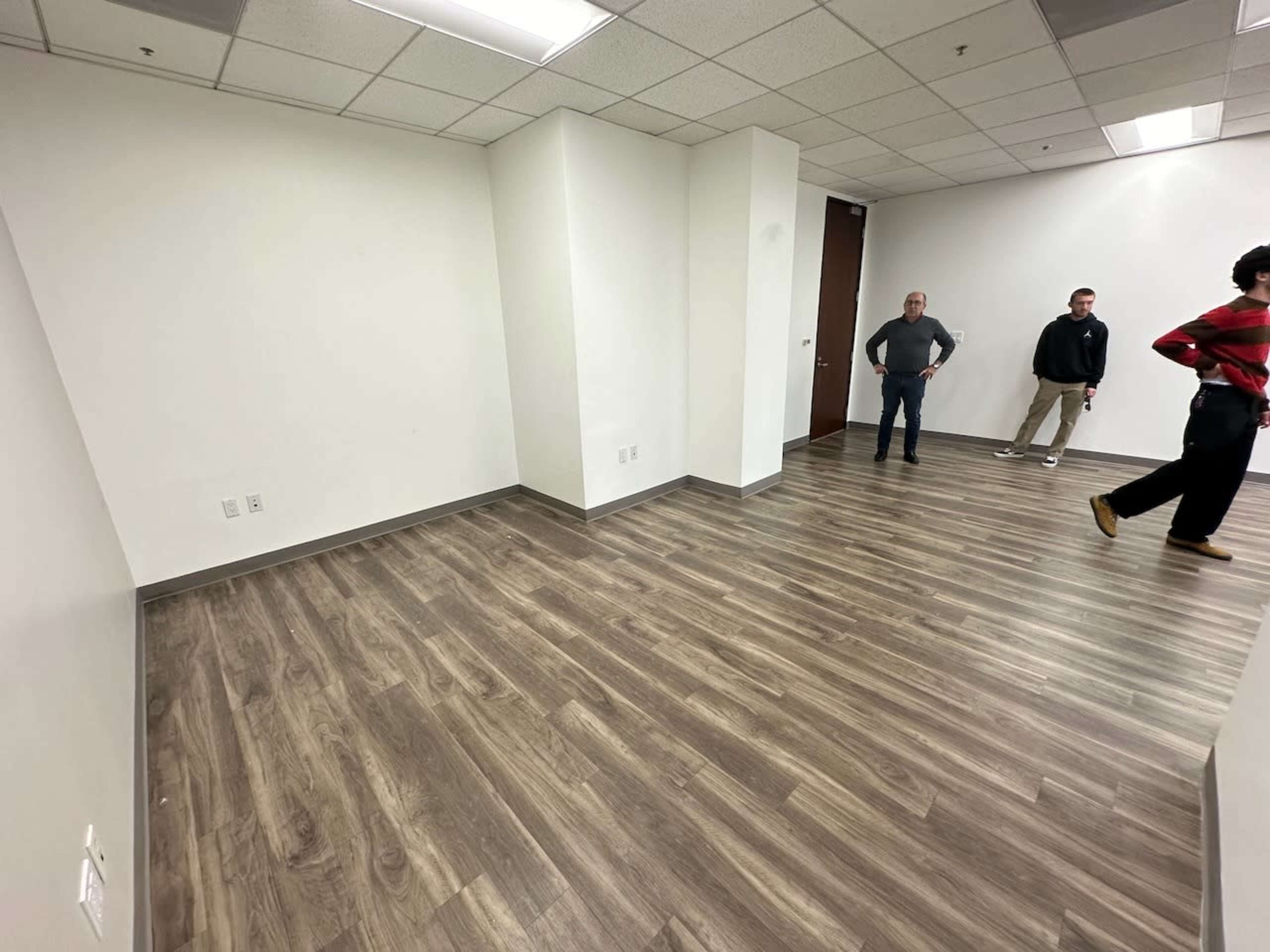 An empty office room with hardwood flooring, two men standing in the background, and a doorway visible on the right.