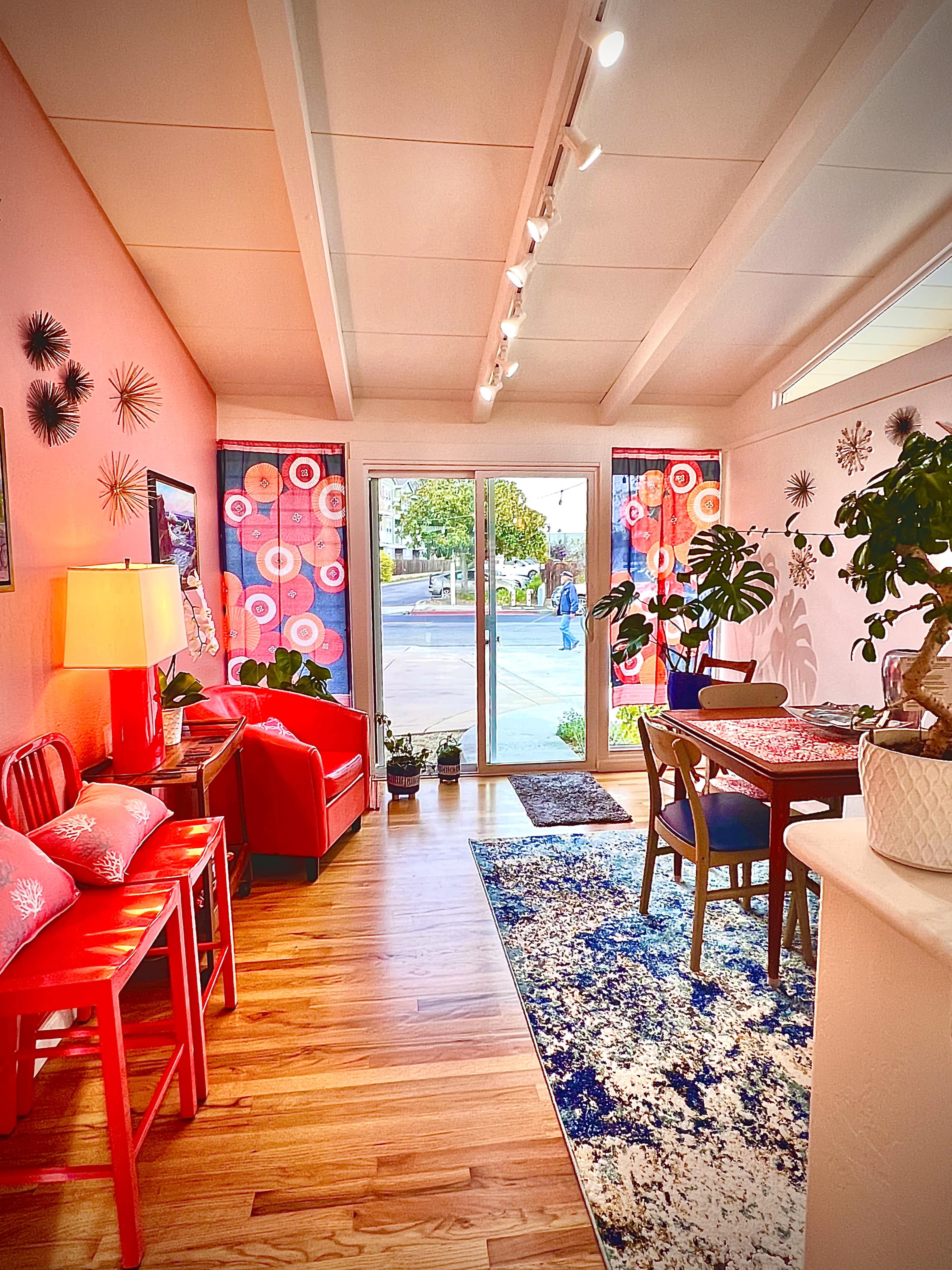 A brightly lit living space featuring a red sofa and chairs, a dining table, and large windows opening to an outdoor view.