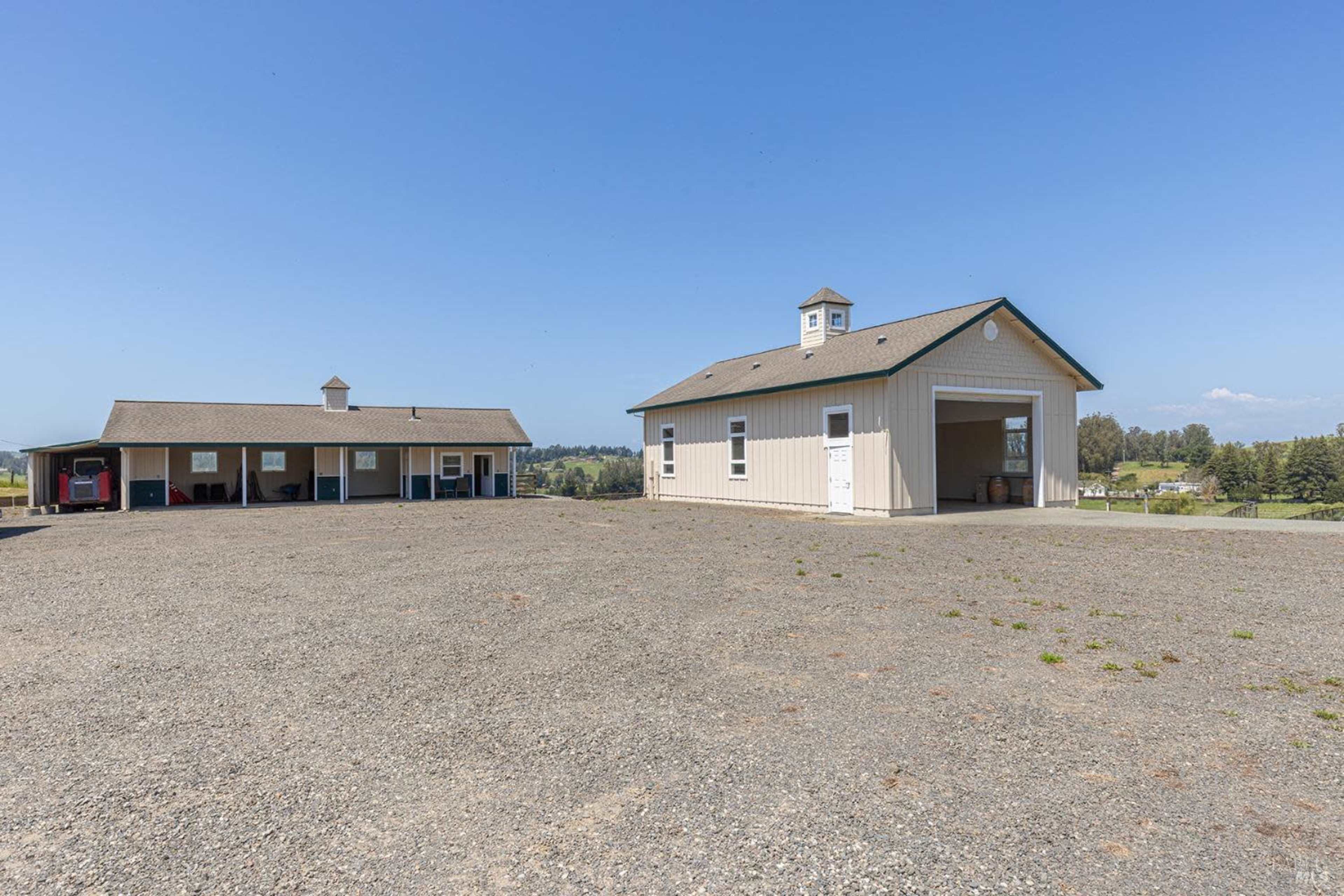 The image shows a large gravel area with two buildings: a garage on the left and a workshop or storage building on the right, set against an open landscape.