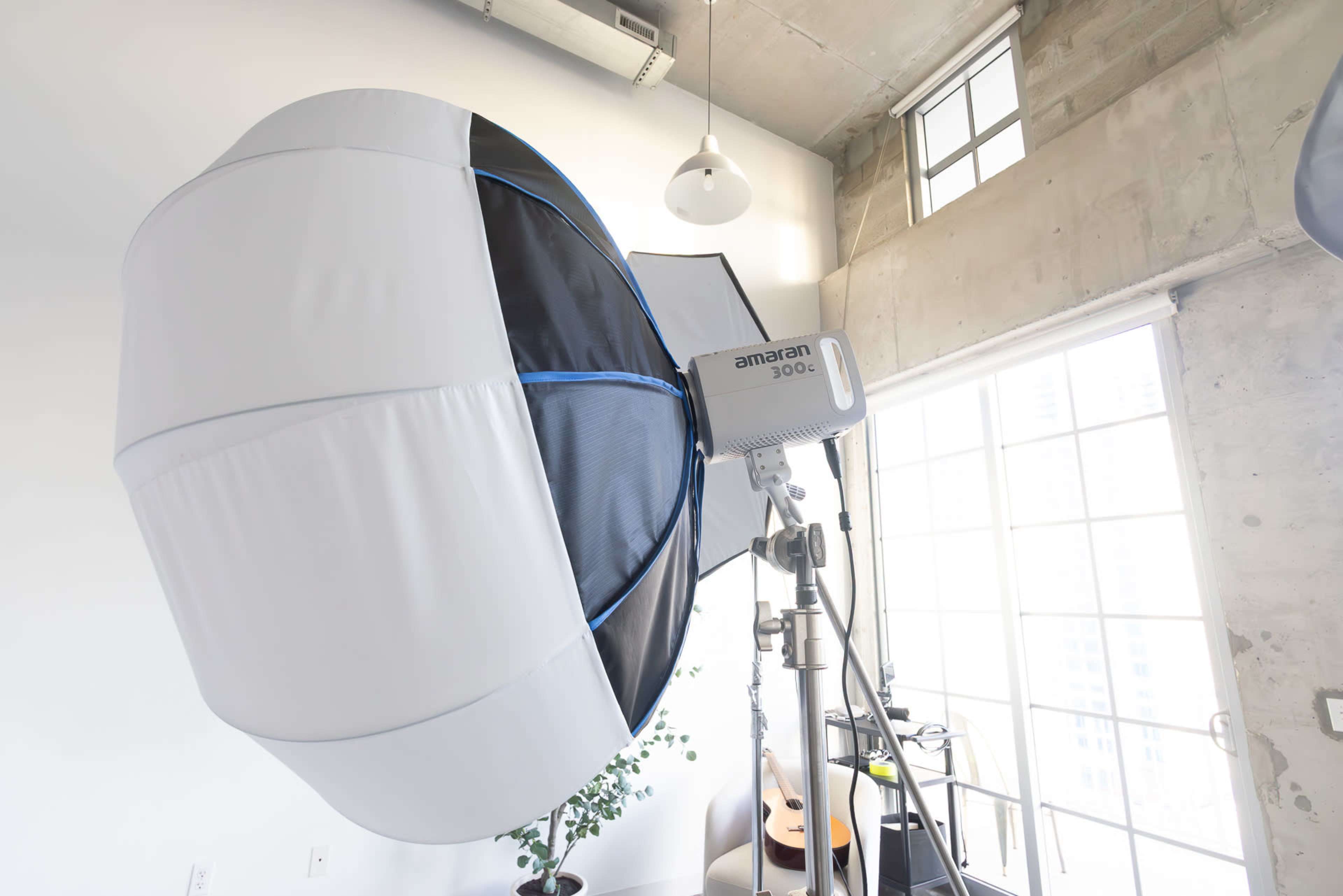 A large softbox light is positioned on a stand in a bright room with large windows and a few indoor plants.