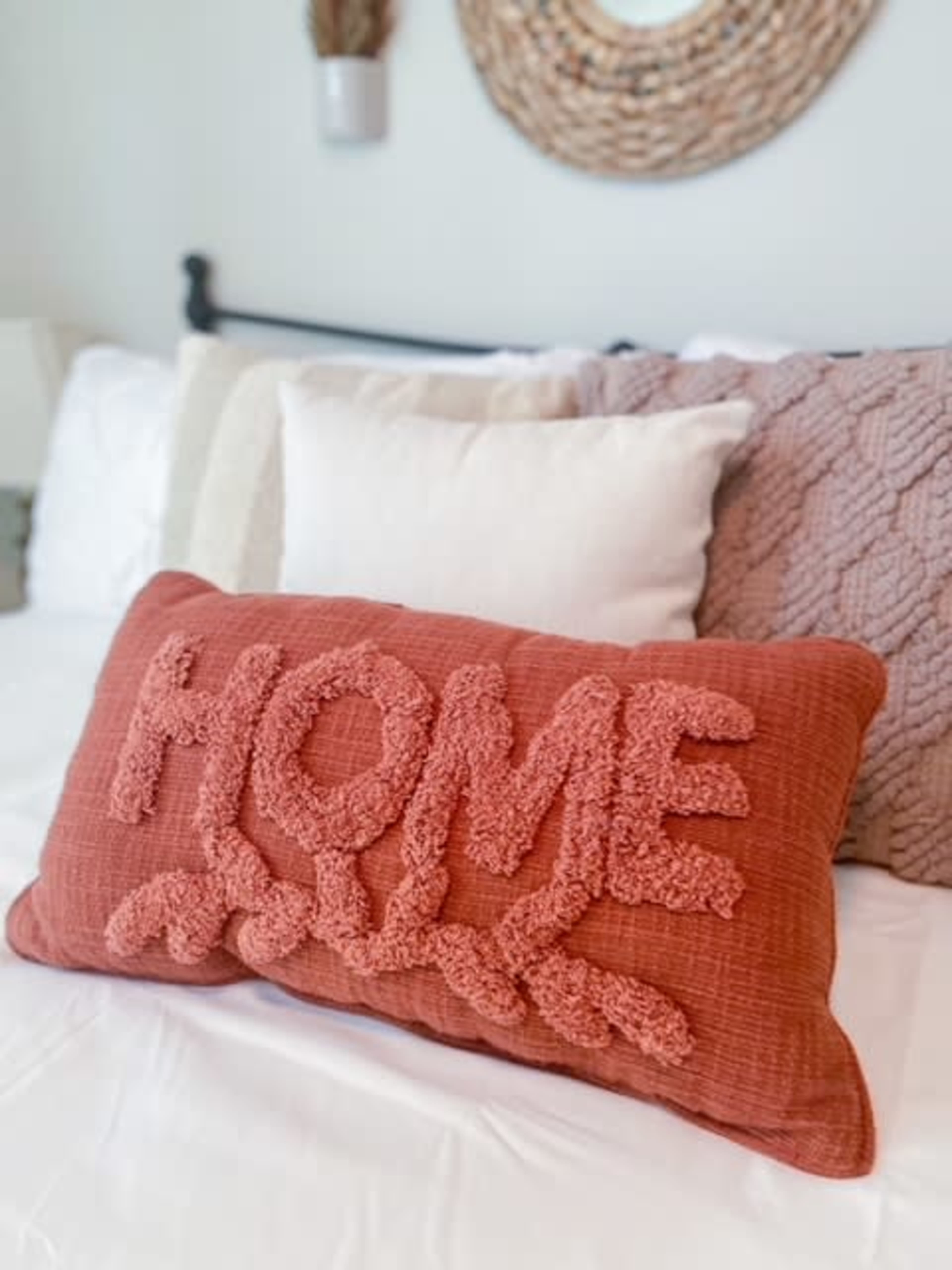 A large, textured pillow with the word "HOME" prominently displayed sits on a neatly made bed adorned with various other decorative pillows.